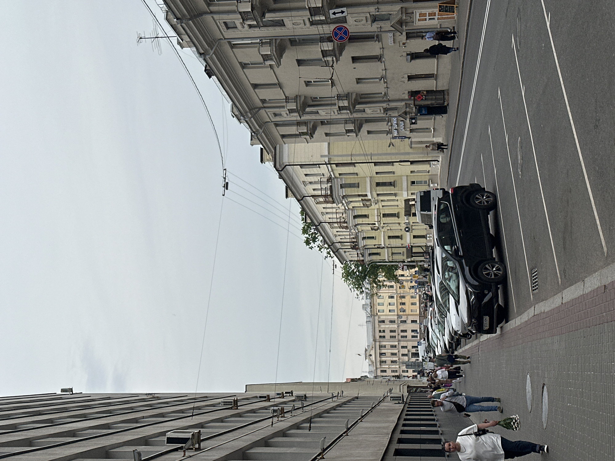 This image depicts a street scene on Valadarskaha street in Minsk, Belarus, under an overcast sky. The view is generally southwest.
On the left, a multi-story, modern grey building, part of the National Bank of the Republic of Belarus complex, features a grid-like facade with vertical and horizontal indentations and several external air conditioning units. A paved sidewalk runs alongside this building, where approximately seven pedestrians are observed. In the foreground, a woman wearing a white t-shirt and dark trousers walks towards the viewer, holding a bouquet of white flowers.
On the right side of the street, a row of older, multi-story buildings with cream-colored facades features balconies and decorative architectural elements. Shopfronts are visible at street level; one sign explicitly reads "РЕМОНТ СМАРТФОНОВ" (Smartphone Repair) and "БАРБЕРШОП" (Barbershop), while another partially visible sign may indicate "КОЛБАСЫ" (Sausages). Two individuals are walking on the sidewalk towards the background on this side.
The asphalt street separates the two sides, featuring marked white lines for parking spaces. Several cars are parked along the right curb, including a black SUV with the Belarusian license plate "9731 KH-7." Overhead utility wires crisscross the upper portion of the frame. A red circular "No Stopping" sign with a blue 'X' and a vertical double-headed arrow is mounted on a pole on the right side of the street.
In the distant background, more traditional buildings are visible, including a prominent light-colored building with a classical facade and a small flag on its roof, which is part of the architectural complex surrounding Independence Square, likely the House of Government.