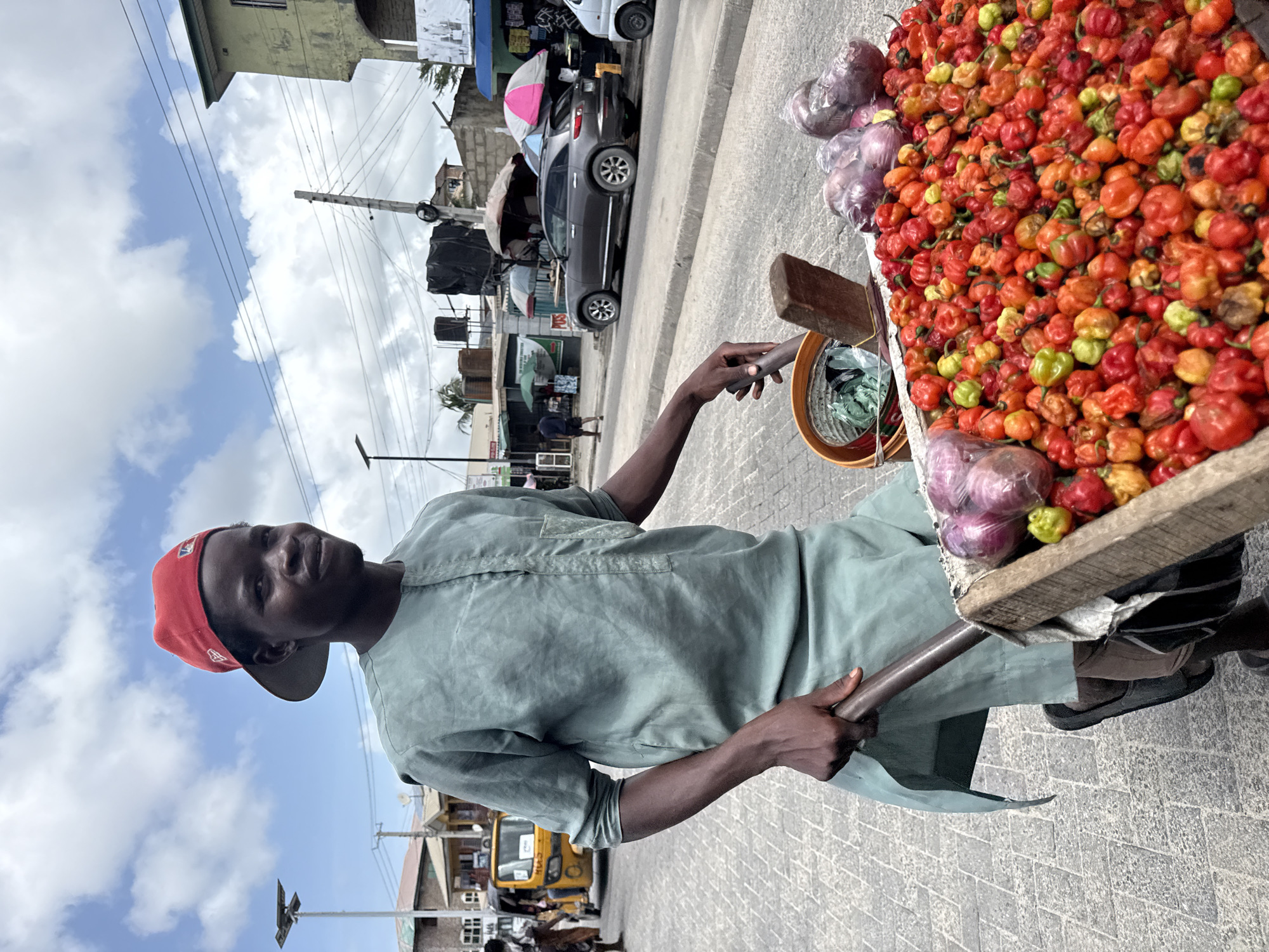 A man with dark skin and a red baseball cap is pulling a cart filled with peppers and onions in what appears to be the city of Ado, Nigeria. He is wearing a light green tunic. The cart is wooden, and the peppers are a mix of red, orange, and green. Some onions, wrapped in plastic, are also visible. The street is paved with gray bricks. In the background, there are buildings, vehicles, and other people. The sky is blue with scattered clouds.
