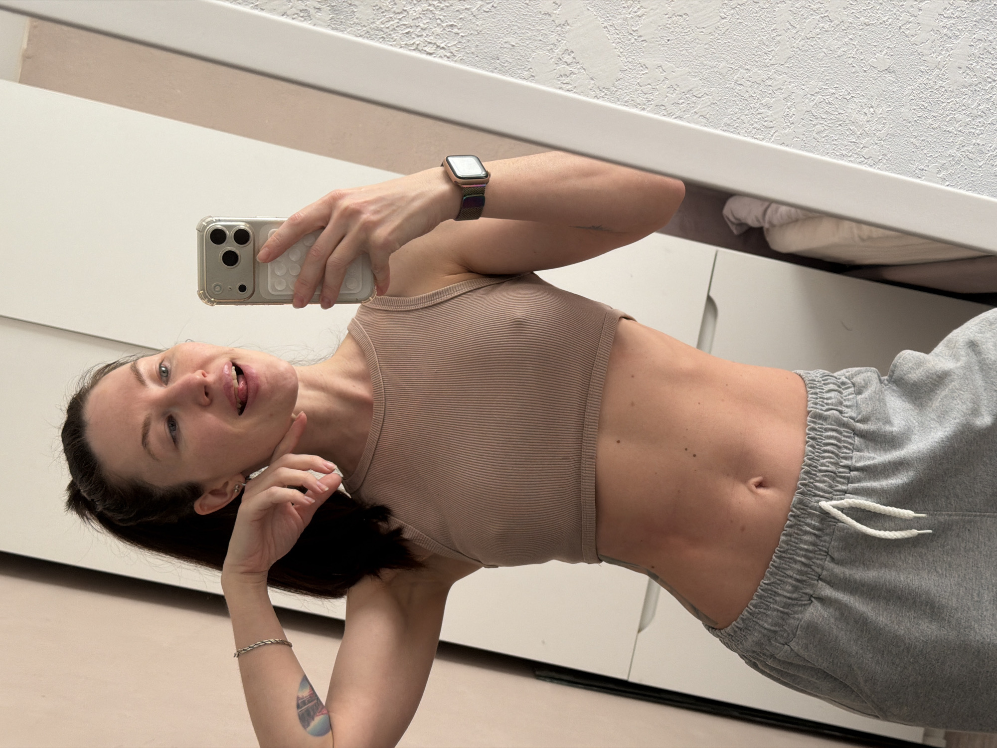 A woman is holding a smartphone and taking a selfie in a mirror. She is wearing a tan-colored tank top and gray sweatpants. The background appears to be an interior with white furniture and white walls. There is no indication that the image was taken in Perm, Russia.