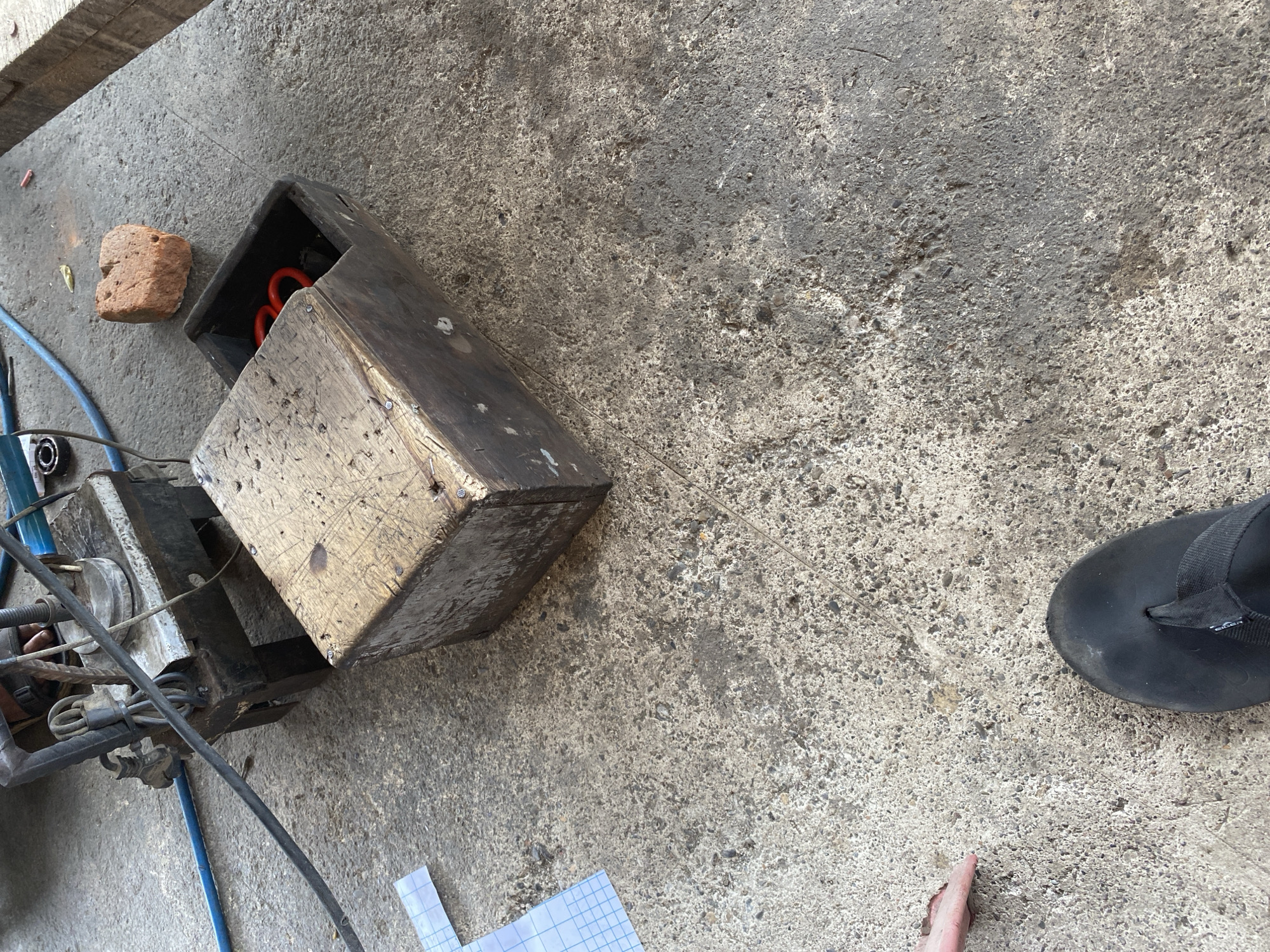 This is a medium-angle, overhead shot of a rough concrete surface, likely outdoors. The concrete is a mix of gray tones with visible aggregate and some staining. In the upper left quadrant of the image, a metal contraption with wires and blue tubing is partially visible. To the right of this, a wooden box with a metal top and a hinged lid is prominent. Inside the open lid of the box, two red, circular objects are visible. Nearby, a single, reddish-brown brick rests on the concrete. Towards the bottom of the image, a left foot wearing a black flip-flop is visible, and a human hand with visible skin texture is reaching towards the center of the frame. A piece of paper with a grid pattern is lying on the concrete near the contraption. The overall environment suggests a workshop or outdoor storage area, possibly in Bayu, Indonesia, given the context provided.