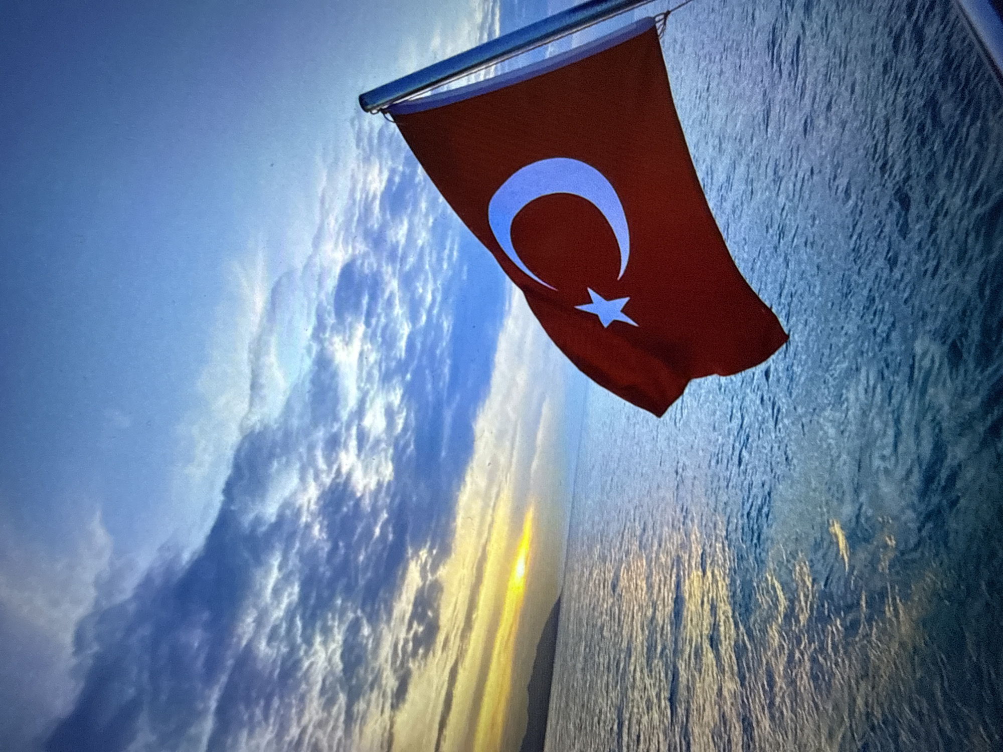 A Turkish flag, red with a white crescent and star, waves from a pole on the right side of the frame, angled towards the left. The flag is in the foreground, obscuring a portion of the ocean in the lower right corner. The ocean, rendered in shades of blue, fills the bottom half of the image, showing subtle ripples and reflections of light. In the background, a low, dark landmass is visible on the left horizon, silhouetted against a bright, hazy yellow and orange sunset. The sky above is a gradient of deep blue, transitioning to lighter blues and whites, with scattered, textured clouds that catch the warm light of the setting sun. The overall scene depicts a serene seascape with a national flag against a dramatic sunset sky.