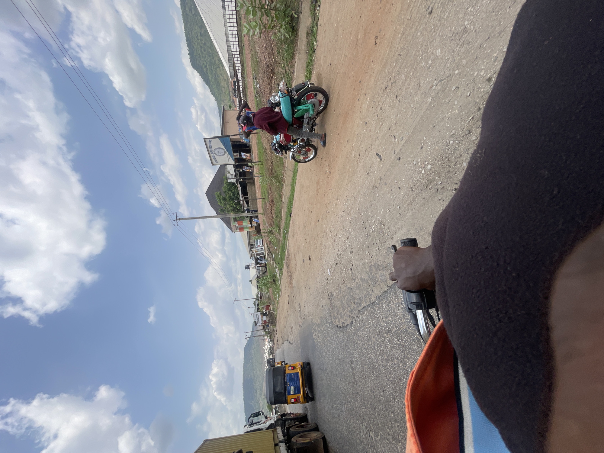 The image depicts a street scene in Lokoja, Nigeria, taken from the perspective of someone riding a motorcycle. The view is oriented with the left side pointing upwards.
In the foreground, a person's arm and torso wearing a dark jacket with orange and blue stripes are visible, holding the handlebars of a motorcycle. The road stretches out ahead with vehicles, including a yellow tricycle, parked on the side. A truck is partially visible in the lower left.
On the left side of the road, there are buildings, signage, and utility poles. A sign for "Grace Kingdom Redemption Church" is visible. A second motorcycle with two people on it is parked on the right side of the road further ahead. In the background, a hill or mountain is visible. The sky is blue with scattered clouds.