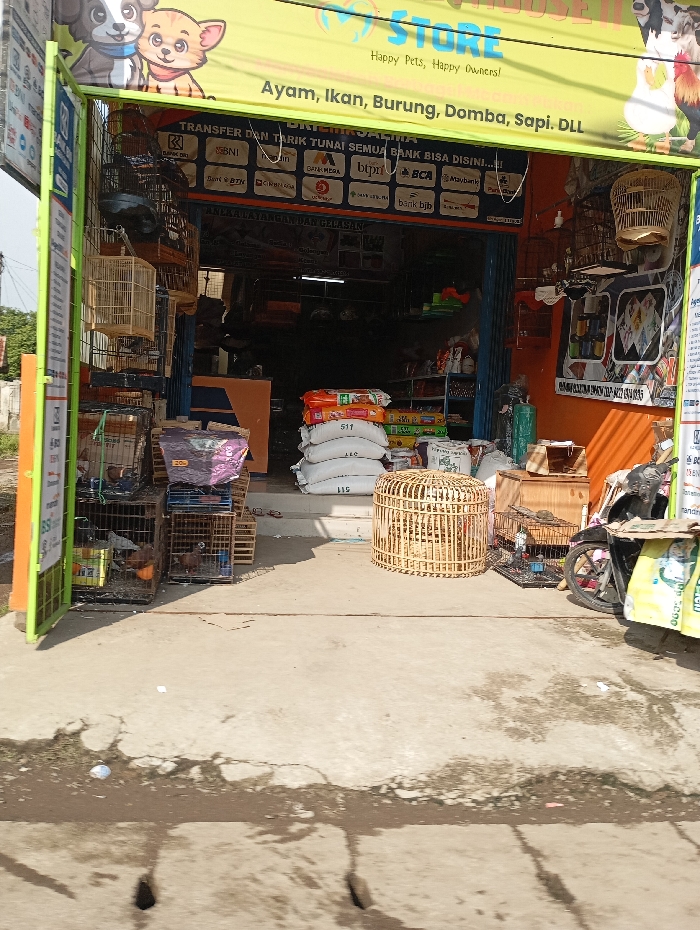 The image displays the exterior of an animal feed and supply store in Cipatik, Indonesia, captured during daylight. The establishment is fronted by a yellow banner with "STORE" prominently displayed, advertising "Happy Pets, Happy Owners!" and services for "Ayam, Ikan, Burung, Domba, Sapi. DLL" (Chicken, Fish, Birds, Sheep, Cattle. Etc.). A secondary banner beneath indicates financial services with multiple bank logos and the text "TRANSFER DAN TARIK TUNAI SEMUA BANK BISA DISINI!!!" (Transfer and withdraw cash from all banks can be done here!!!).
The store entrance features a partially open dark blue metal roll-up door, revealing stacks of white bags (likely feed, one marked "511") and other merchandise within. The right exterior wall is orange, while the left door frame is green.
Outside the store, several animal cages are visible. Multiple wooden bird cages hang from the left side of the entrance frame. On the ground to the left, several wire cages contain birds, including what appear to be chickens or pigeons. A large, round, light-colored wicker or bamboo cage rests on the ground directly in front of the entrance. A black motorcycle is parked to the right, adorned with a yellow and green bag.
The foreground consists of cracked concrete pavement with scattered small debris and a dark, damp-looking patch. Drainage holes are visible at the bottom of the frame, indicating a street or sidewalk environment. No individuals are present in the scene, and no actions or interactions are depicted.