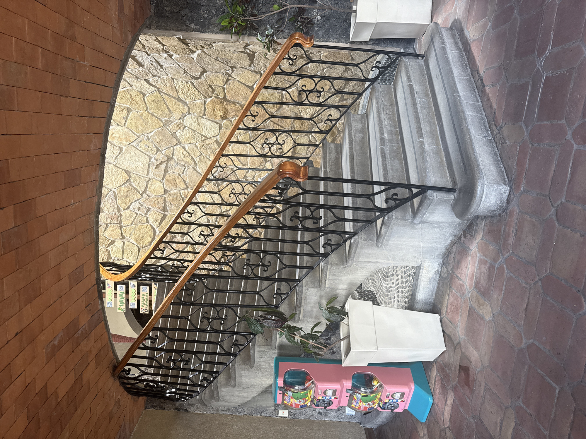 The image depicts an indoor staircase with a unique architectural design. The staircase has a wooden handrail with black metalwork that features decorative swirls and a repeating pattern. The steps are made of grey stone and are flanked by walls of contrasting materials. One wall is made of red bricks, while the other is made of tan-colored stone blocks. The staircase appears to be in Tres de Mayo, Mexico. At the bottom of the staircase, there is a pink candy machine with multiple compartments. A white rectangular object is also present near the candy machine. A plant is situated near the steps, adding a touch of greenery to the scene. The flooring around the candy machine is also made of stone.