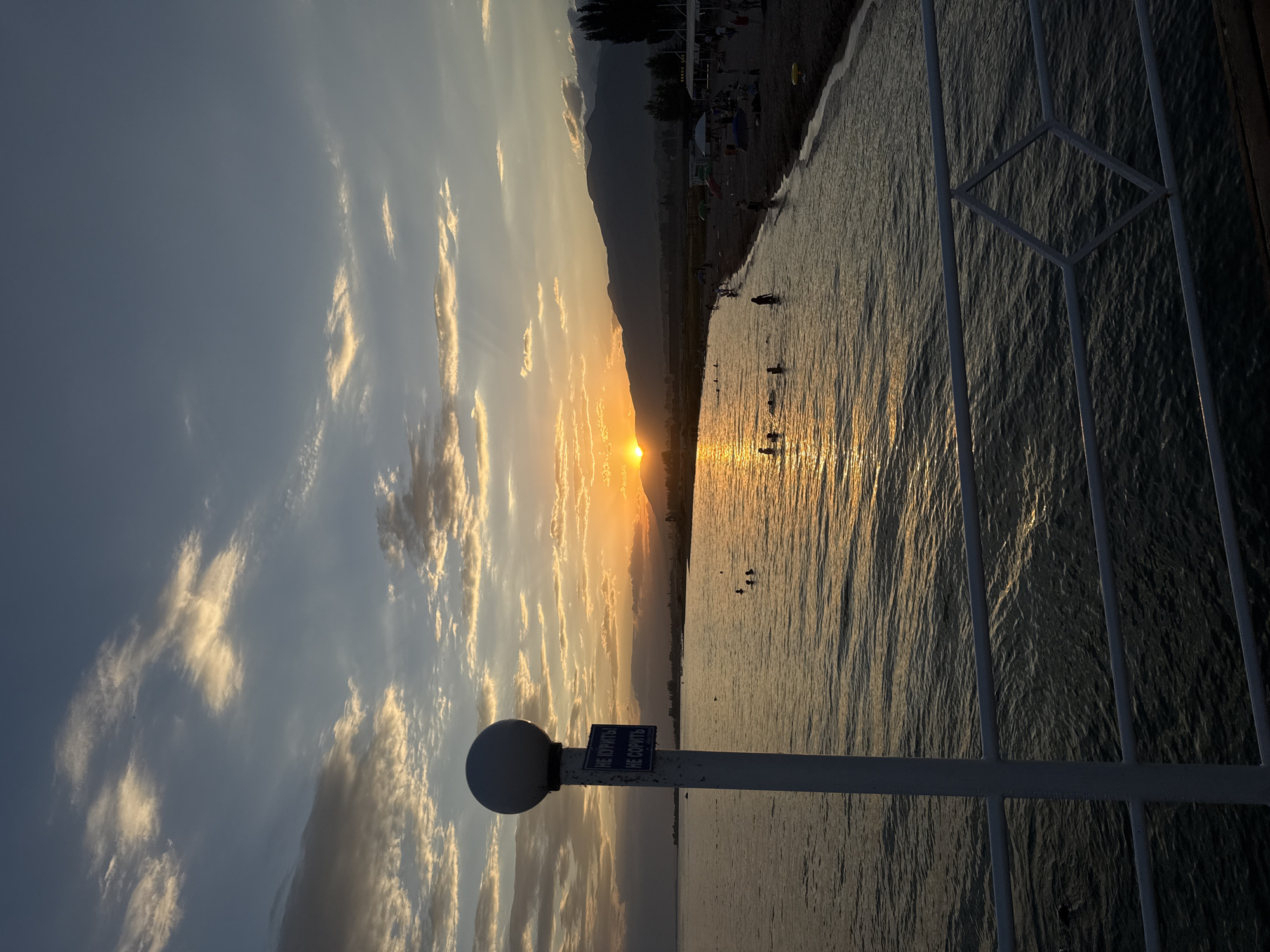 The image captures a sunset over Issyk-Kul Lake near Sary-Oy, Kyrgyzstan. The sky is filled with a mix of blue and orange hues, with scattered clouds illuminated by the setting sun. The sun is low on the horizon, casting a golden reflection on the water. On the lake, several people are swimming or wading near the shore.
The foreground features a white railing and a lamppost with a sign attached. The sign has text in a Cyrillic alphabet, suggesting a Russian or Kyrgyz inscription. Beyond the water, a sandy beach is visible, populated by a few individuals and some beach umbrellas. In the background, silhouetted mountains rise against the colorful sky. The overall scene depicts a tranquil evening at a lakeside resort.