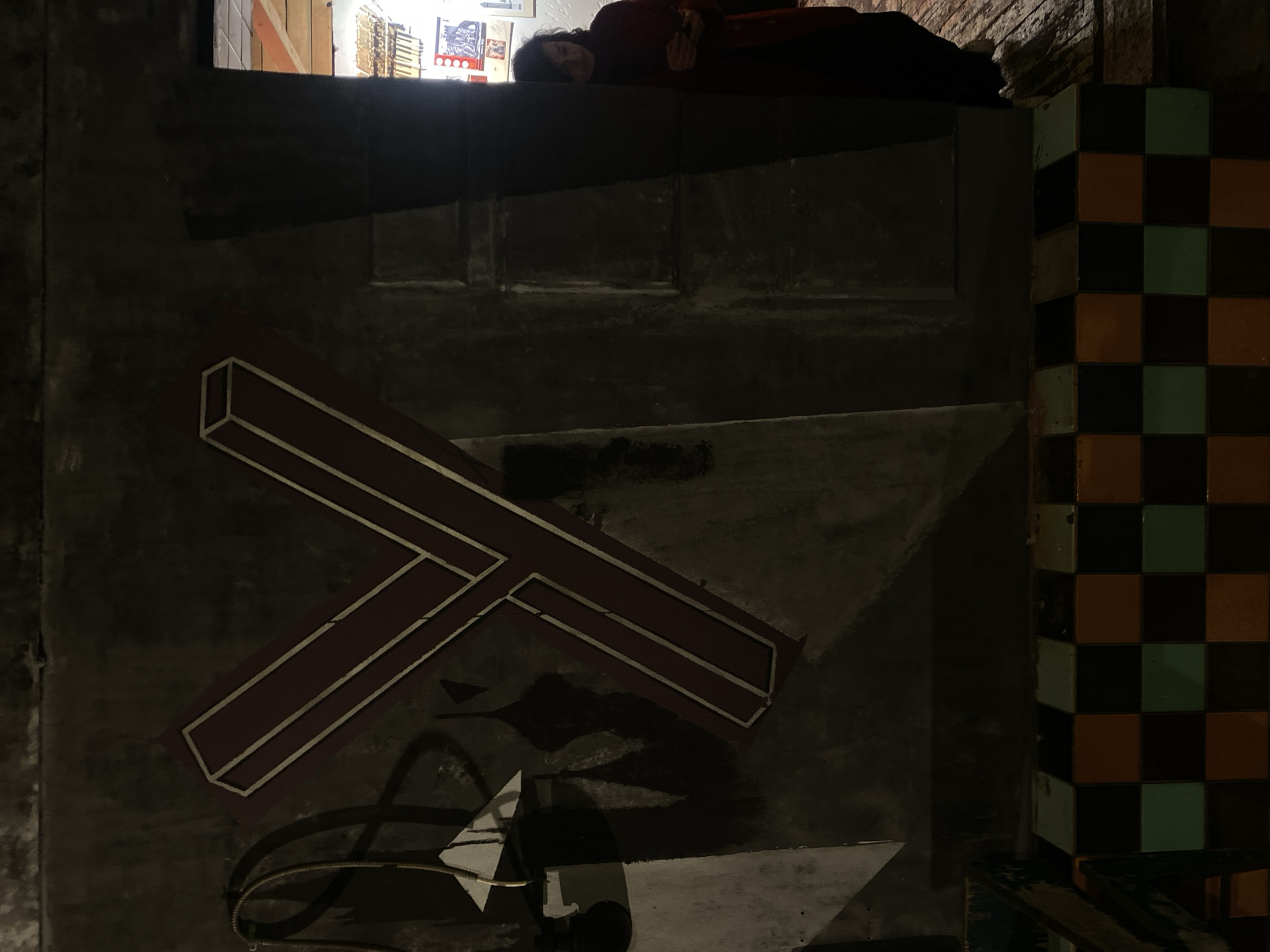 The image depicts an urban scene, possibly a dimly lit alley or underpass in Moscow, Russia. A woman is lying on an elevated ledge at the top of the frame, appearing to be resting or sleeping. Below her, the wall is adorned with abstract graffiti, including geometric shapes, lines, and shadows. A tiled column with a checkerboard pattern in shades of brown, green, and black is located on the right side of the frame. The overall ambiance is dark and somewhat gritty, suggesting an unconventional or neglected space.