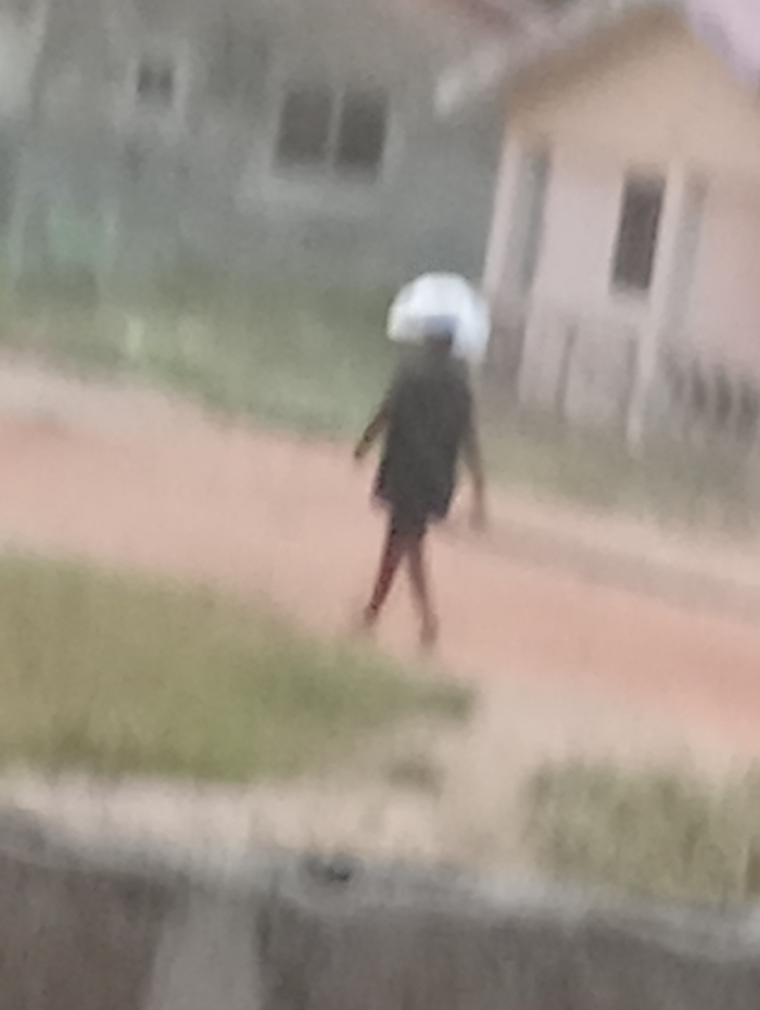 A blurred image shows a person walking away from the camera on a dirt road in what appears to be a residential area. The person is wearing a dark shirt and carrying something white on their head. In the background are buildings with windows. Green vegetation is visible near the road. The location is Unknown, Unknown.