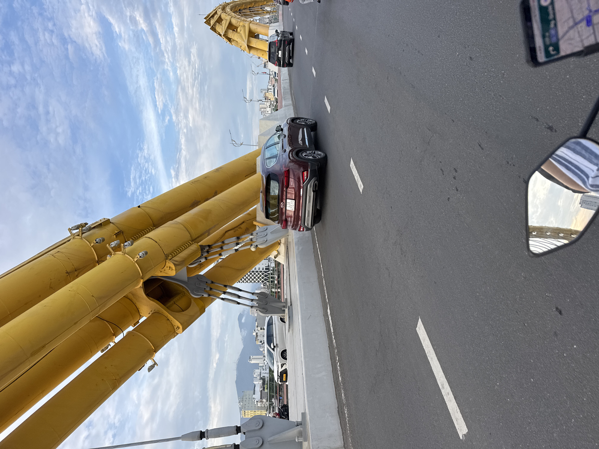 The image captures a view from the road deck of the Dragon Bridge in Da Nang, Vietnam. Dominating the frame are the massive, bright yellow steel tubes of the bridge structure, suspended by thick cables. The road surface is asphalt with white lane markings. Several vehicles are visible on the bridge: a maroon Hyundai Santa Fe SUV is in the foreground, a black SUV is further ahead in the right lane, and a white sedan is on the left side of the bridge, partially obscured by the yellow structure. In the background, the cityscape of Da Nang unfolds, with buildings and mountains under a cloudy sky. A portion of a motorcycle's rearview mirror and handlebars are in the bottom right and bottom left corners, indicating the vantage point is from a motorcycle.
