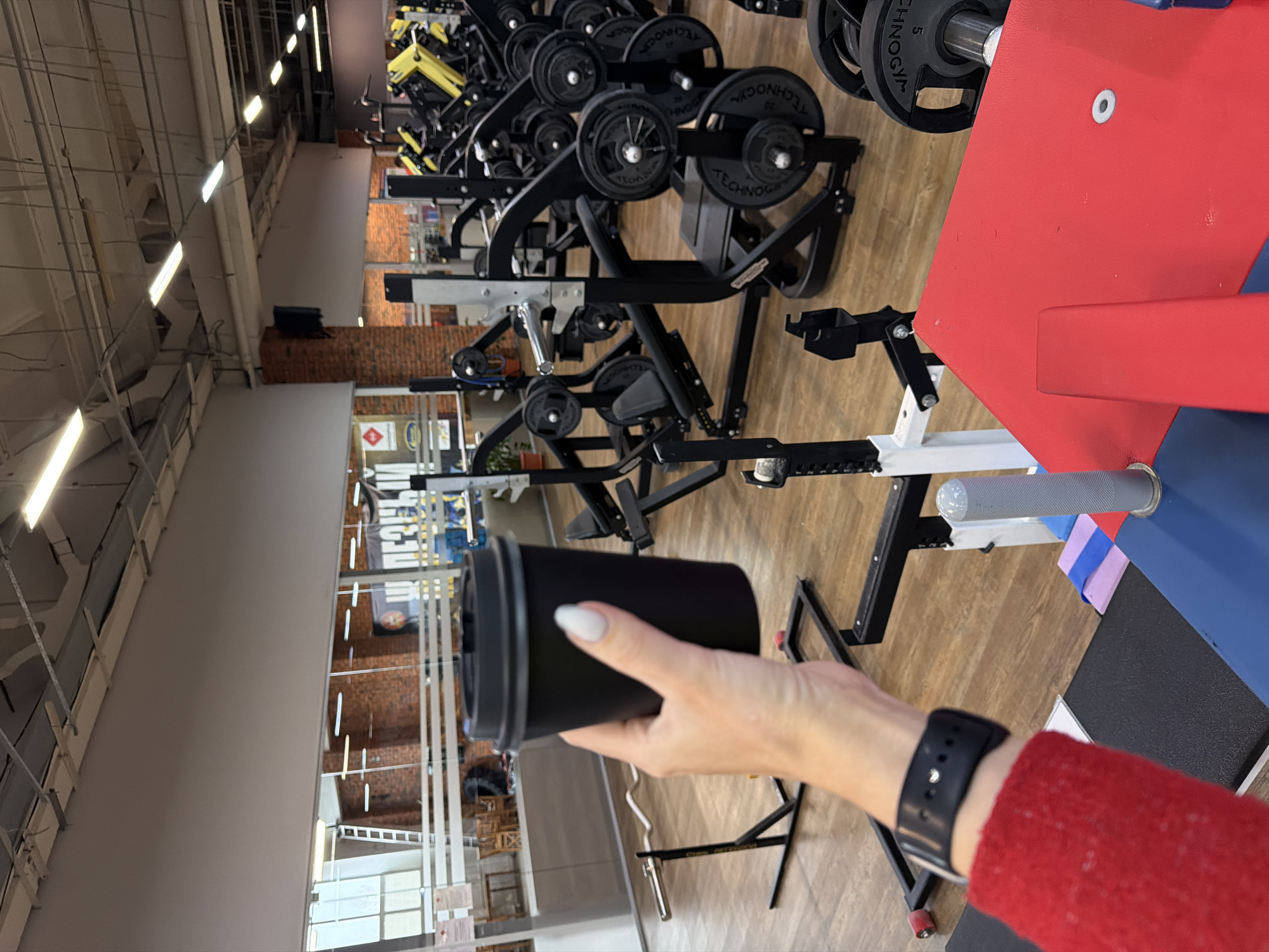 A person holds a black coffee cup in a gym in Chaikovsky, Russia. The person wears a red sweater and a black smartwatch. The gym contains black weightlifting equipment with visible weights. The gym has a wood floor and brick and white walls with overhead fluorescent lights.
