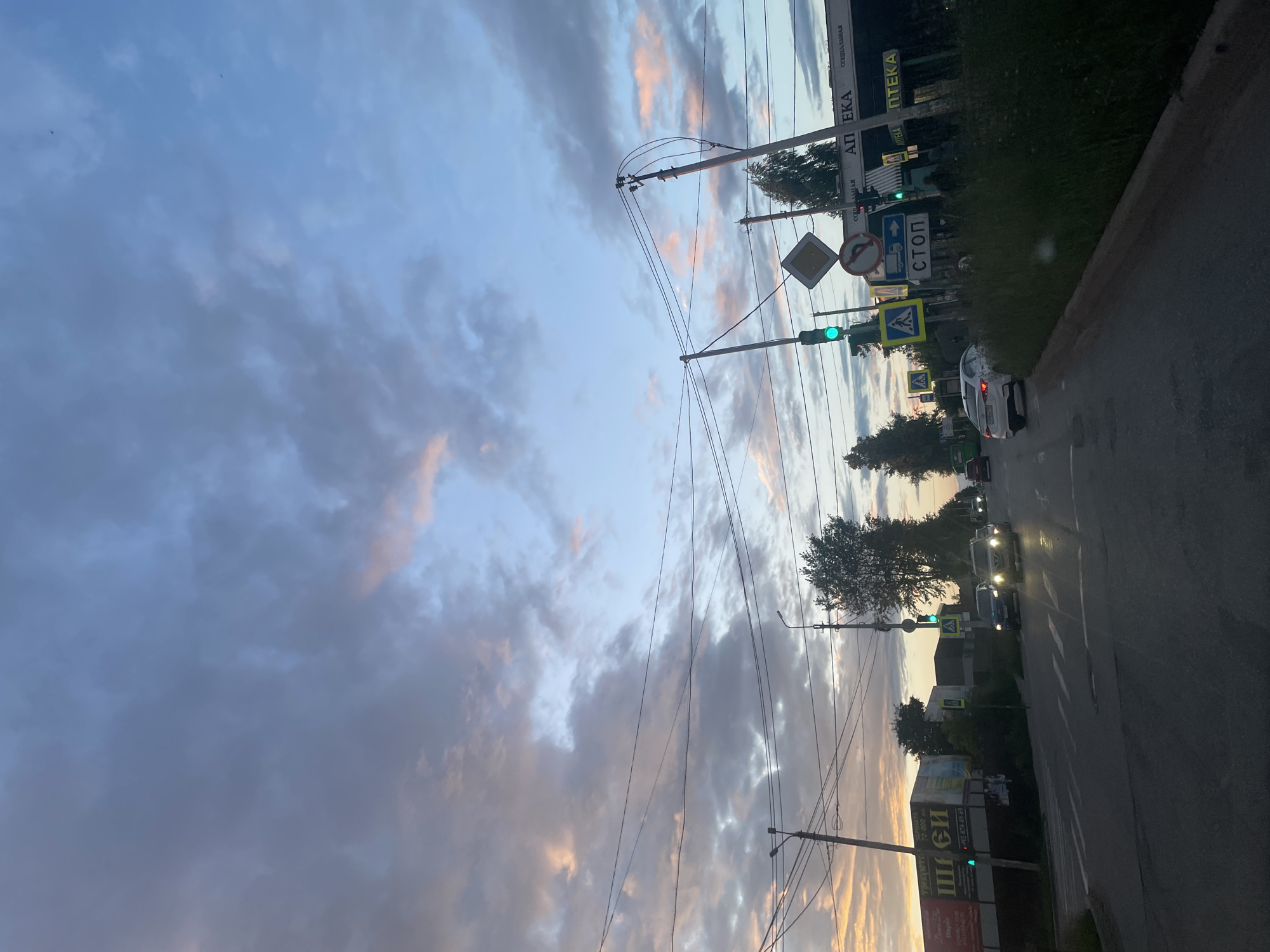 This media file captures a street scene at dusk in Votkinsk, Russia. The image is oriented vertically, with the sky taking up a significant portion of the frame. Clouds, tinged with pink and orange from the setting sun, drift across a pale blue sky. Power lines crisscross the sky, supported by utility poles.
Below the sky, a street runs from the foreground towards the background. Several cars are visible on the road, some with headlights on, suggesting the approaching night. Traffic lights show a green signal, indicating that traffic can proceed. Road signs are present, including a stop sign, a yield sign, and pedestrian crossing signs, all typical of a road in a populated area.
Along the side of the road, there are buildings with signage, one of which appears to be an "АПТЕКА" (Apteka), meaning pharmacy. Other signs are partially visible. Trees line the side of the road, and a dense patch of greenery is also present. The overall impression is a quiet evening on a suburban or small-town street as daylight fades. The lighting and clouds suggest it is either sunrise or sunset, with the sky colors leaning towards sunset.