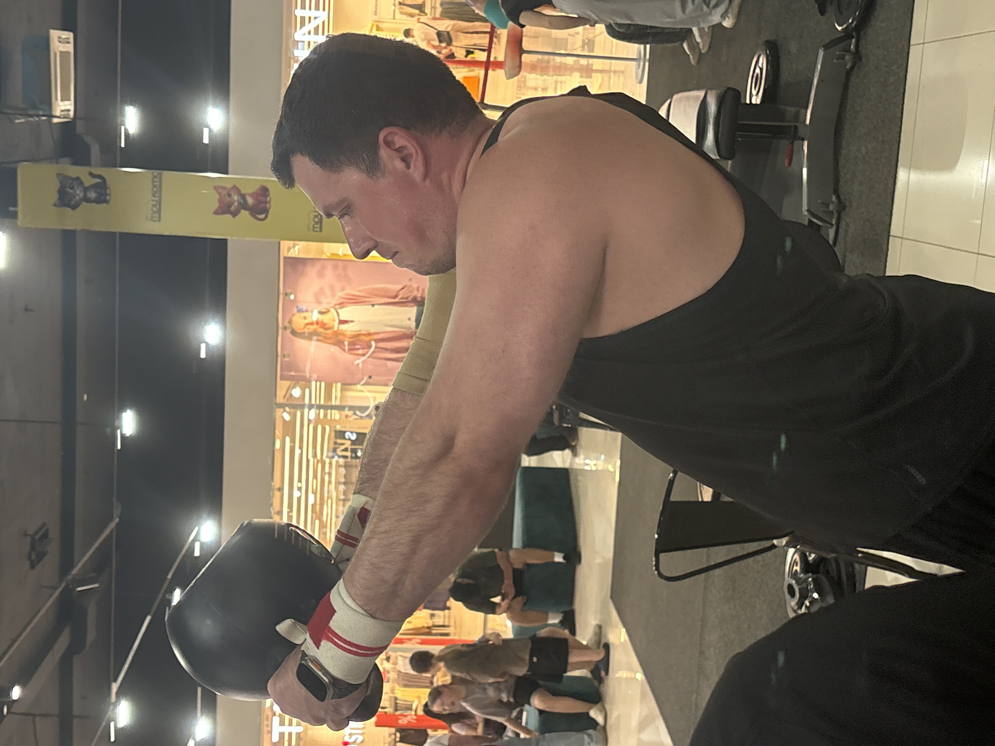 The image shows a man in a black tank top holding a black kettlebell in front of him with both hands. He is wearing red and white wristbands. He is in an indoor setting that appears to be a gym or fitness area, with gym equipment visible in the background, including weight plates and a bench. Other people are present in the background, some seated and some standing. The environment is brightly lit, with ceiling lights visible. A yellow pillar with cat decals and text is also visible in the background. The man's face is visible in profile, and he appears to be mid-exercise.