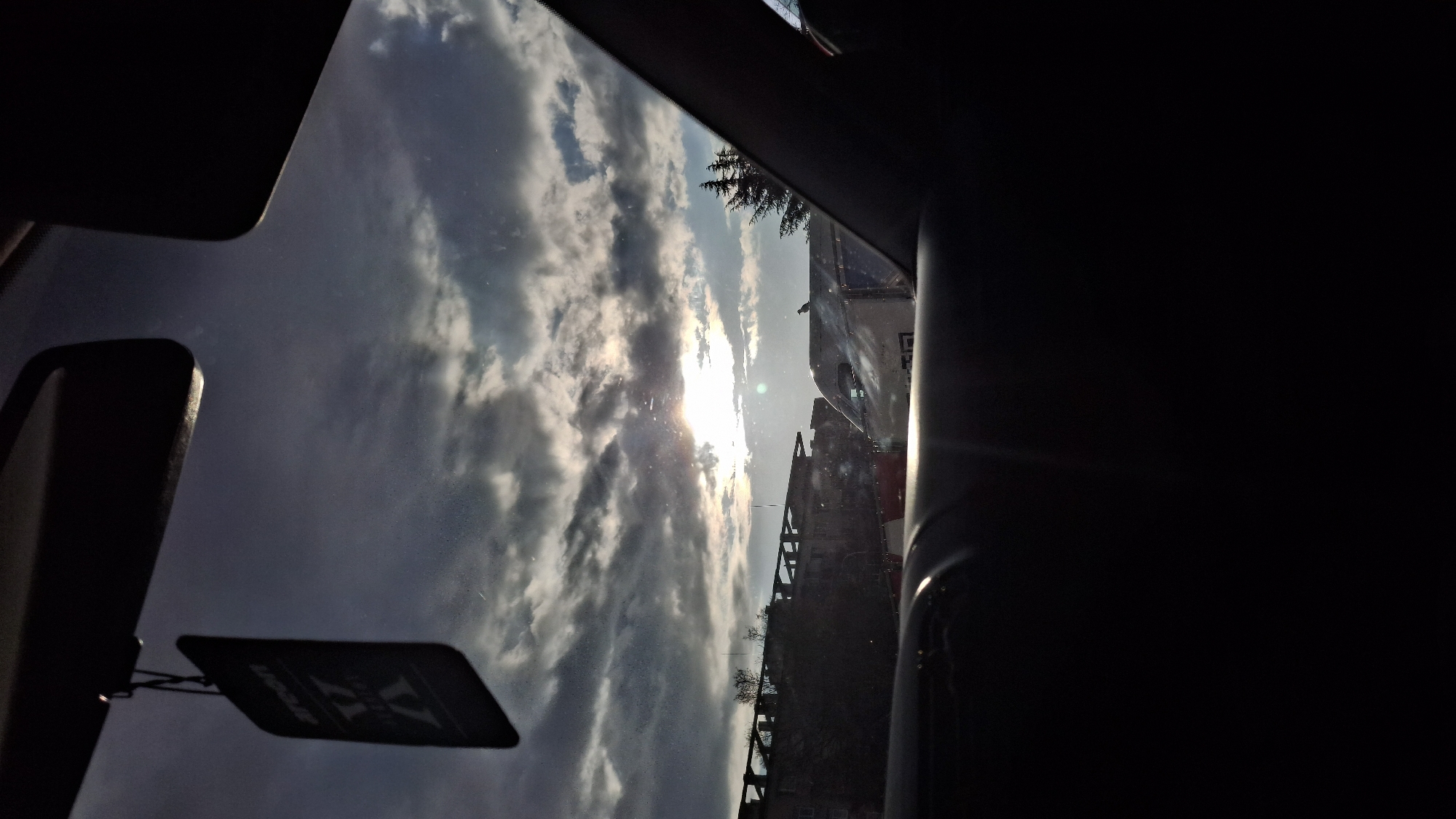The image, captured from inside a vehicle, presents a view through the windshield obstructed by parts of the car's interior, including the rearview mirror and the passenger side pillar. The sky is filled with cloudy, overcast conditions. In the distance, buildings and structures suggest an urban environment. The sun is positioned low in the sky, near the horizon, partially obscured by the clouds, creating a glare on the windshield. The location appears to be the city of Unknown, Unknown, based on the limited information available from the scene.