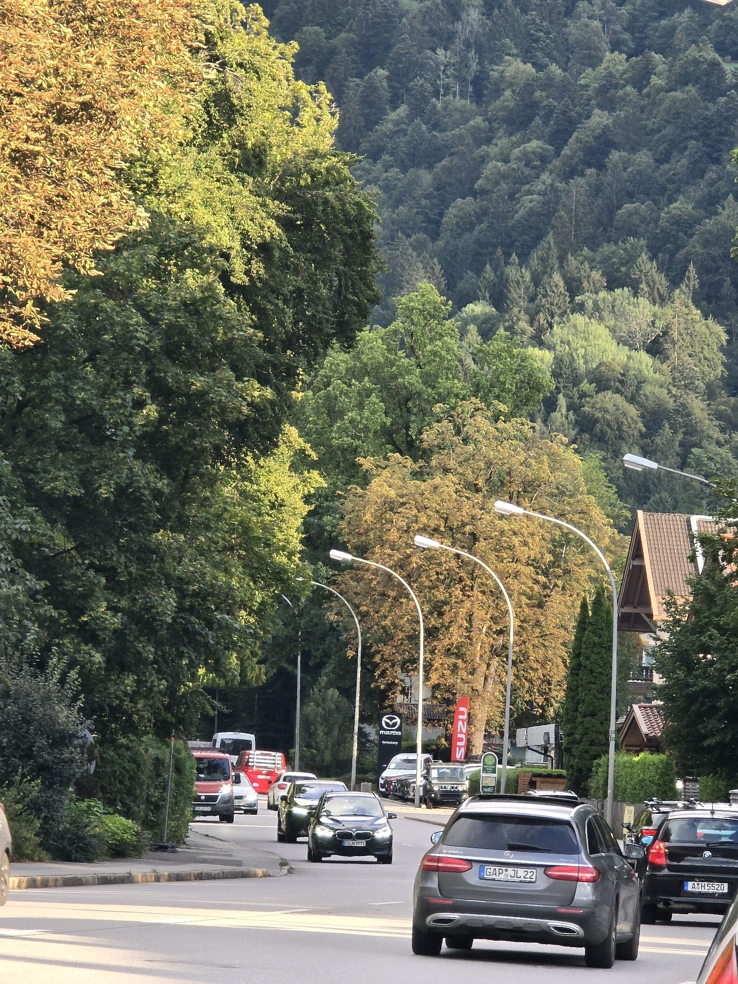 The image depicts a street scene in Garmisch-Partenkirchen, Germany, featuring a traffic-filled road bordered by lush green trees and a wooded hillside. Several cars are visible, including a black Mercedes-Benz E-Class wagon in the foreground with a German license plate ending in "JL 22," and another black Mercedes-Benz sedan with a license plate ending in "H 5520" behind it. Further down the road, a red van and other vehicles are present. Streetlights with curved poles line the street, and a building with a distinctive tiled roof is visible on the right. The background is dominated by a dense forest covering a hillside, with the trees showing a mix of green and autumnal yellow foliage, suggesting it might be late summer or early autumn. The sky is clear, and the sunlight casts shadows, indicating it is daytime.