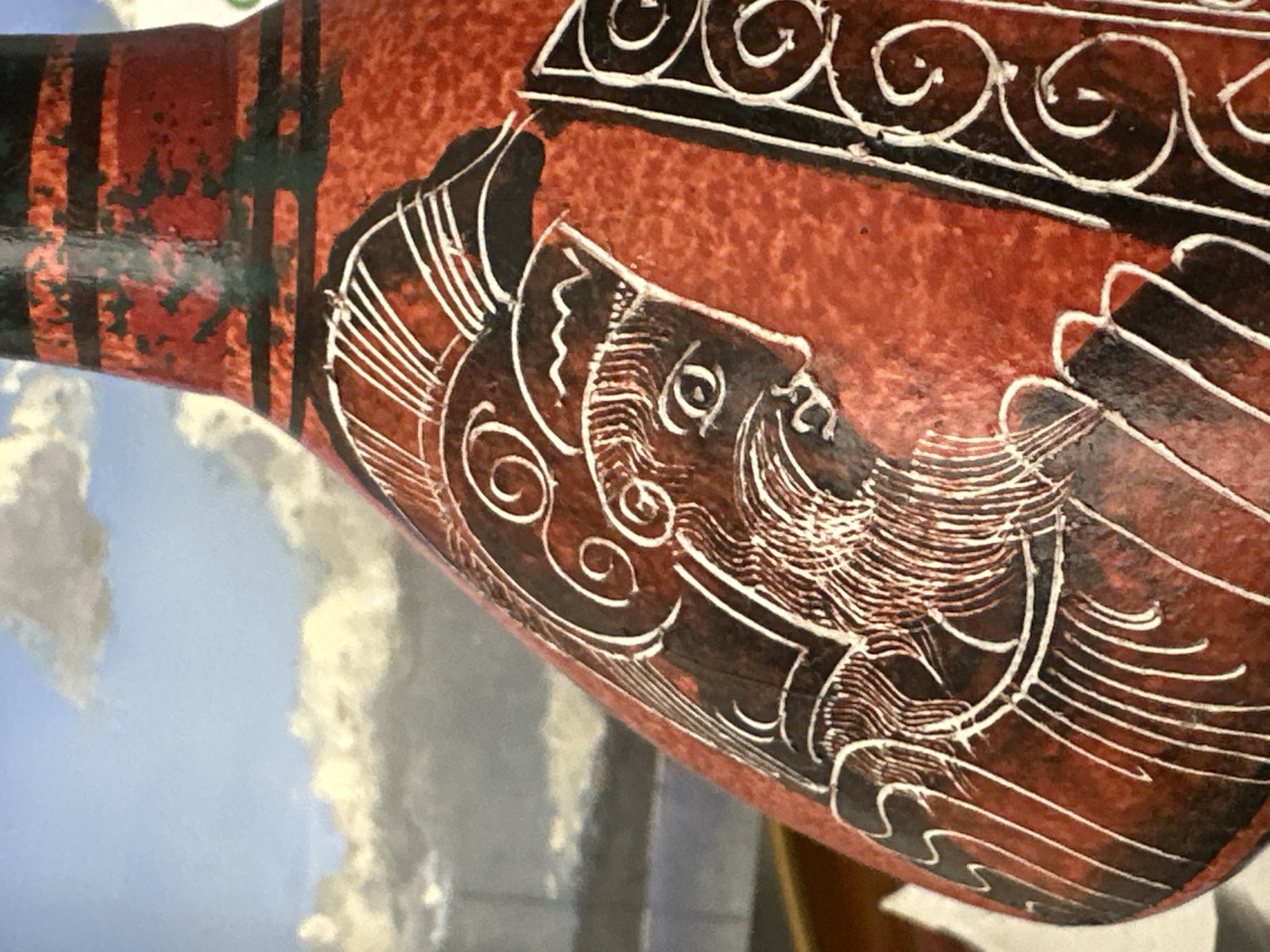 The image shows a close-up of a red, textured vase with a black and white engraved design of a profile of a person. The person has a beard, an eye, and is wearing a helmet. The background appears to be a painted mural of a cloudy sky. There is no indication of the city of Nevinnomyssk, Russia, or any other specific location. The image is a still, isolated shot of the vase.