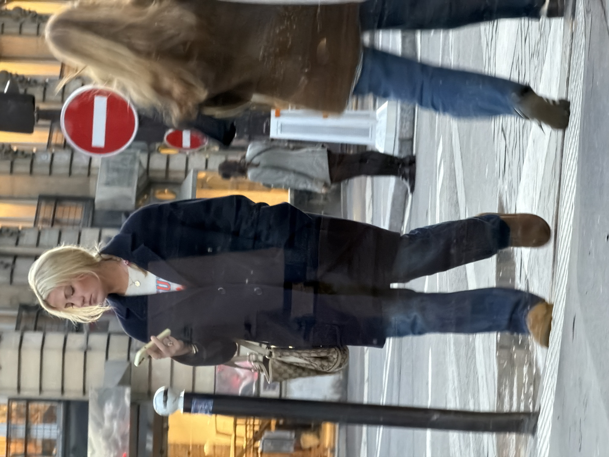The image captures a street scene in Paris, France, featuring three individuals and urban infrastructure.
In the left foreground, a blonde female, appearing to be in her mid-20s to early 30s, walks towards the right. She wears a dark navy long coat over a light-colored t-shirt with a partial red/orange graphic, blue jeans, and light brown suede-like shoes. Her right hand holds a light-colored smartphone, which she is actively viewing, while her left hand is in her coat pocket. She wears a ring on her right ring finger and a gold-toned necklace. A large, light-colored shoulder bag with a visible monogram pattern is draped over her left shoulder. Reflections from an adjacent glass surface are visible across her figure.
To the right of the primary subject, the upper torso and head of a second individual are visible, moving away from the camera. This person has long, wavy blonde or light brown hair and is wearing a brown jacket, blue jeans, and dark shoes. Motion blur is evident on this individual, indicating movement.
In the mid-ground, on the opposite side of the street, a third person is observed from behind. This individual has dark hair pulled back and wears a light grey or beige blazer/jacket over dark trousers, paired with dark tall boots. This person appears to be standing or moving slowly.
The background features multi-story stone buildings characteristic of Parisian architecture, with windows and subtle decorative elements. A prominent red circular 'No Entry' sign (panneau B2a "sens interdit"), with a white horizontal bar, is centrally located in the mid-ground, mounted on a pole, with a smaller identical sign beneath it. The street is paved, displaying white road markings, and the foreground includes a sidewalk with visible tactile paving strips. A black pole stands vertically in the far left foreground, and a white-topped bollard is positioned on the sidewalk in the mid-ground. The lighting suggests either a late afternoon or an overcast day, with some interior lights visible in the background buildings.