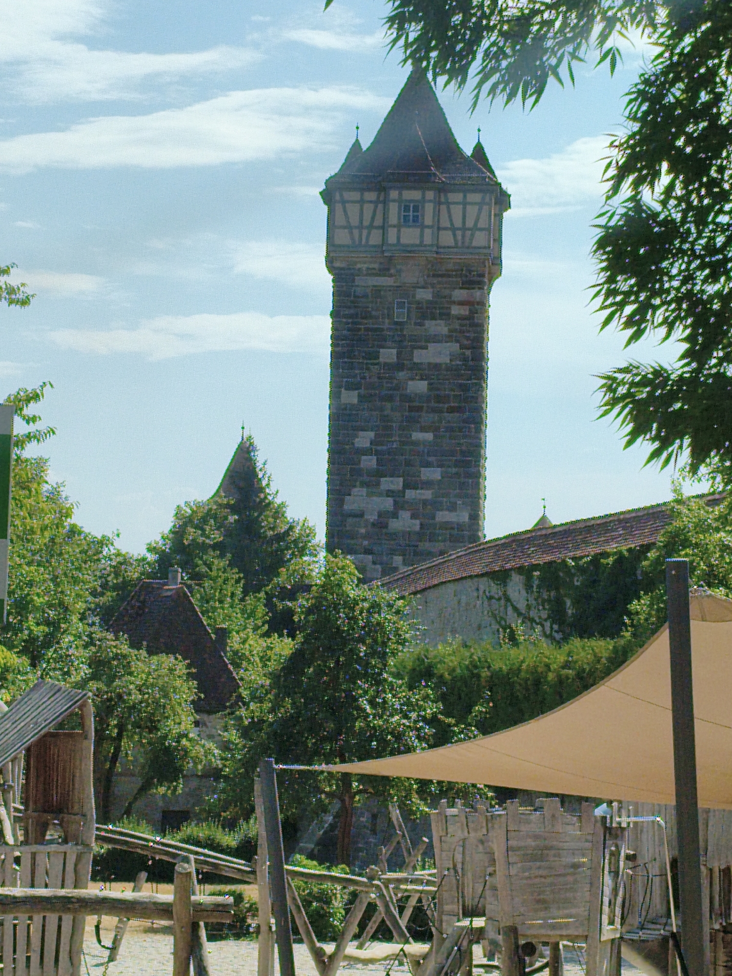 The image shows a stone tower, likely a medieval fortification, rising above a green landscape with trees and buildings. The tower has a distinctive architectural style with a tiled roof and decorative elements. In the foreground, there appears to be a wooden playground structure, possibly for children, with sand or gravel on the ground. A large, beige fabric canopy is stretched over part of the playground, offering shade. The sky is partly cloudy. This scene is likely located in Rothenburg ob der Tauber, Germany, a historic city known for its well-preserved medieval architecture, including its numerous towers and fortifications.