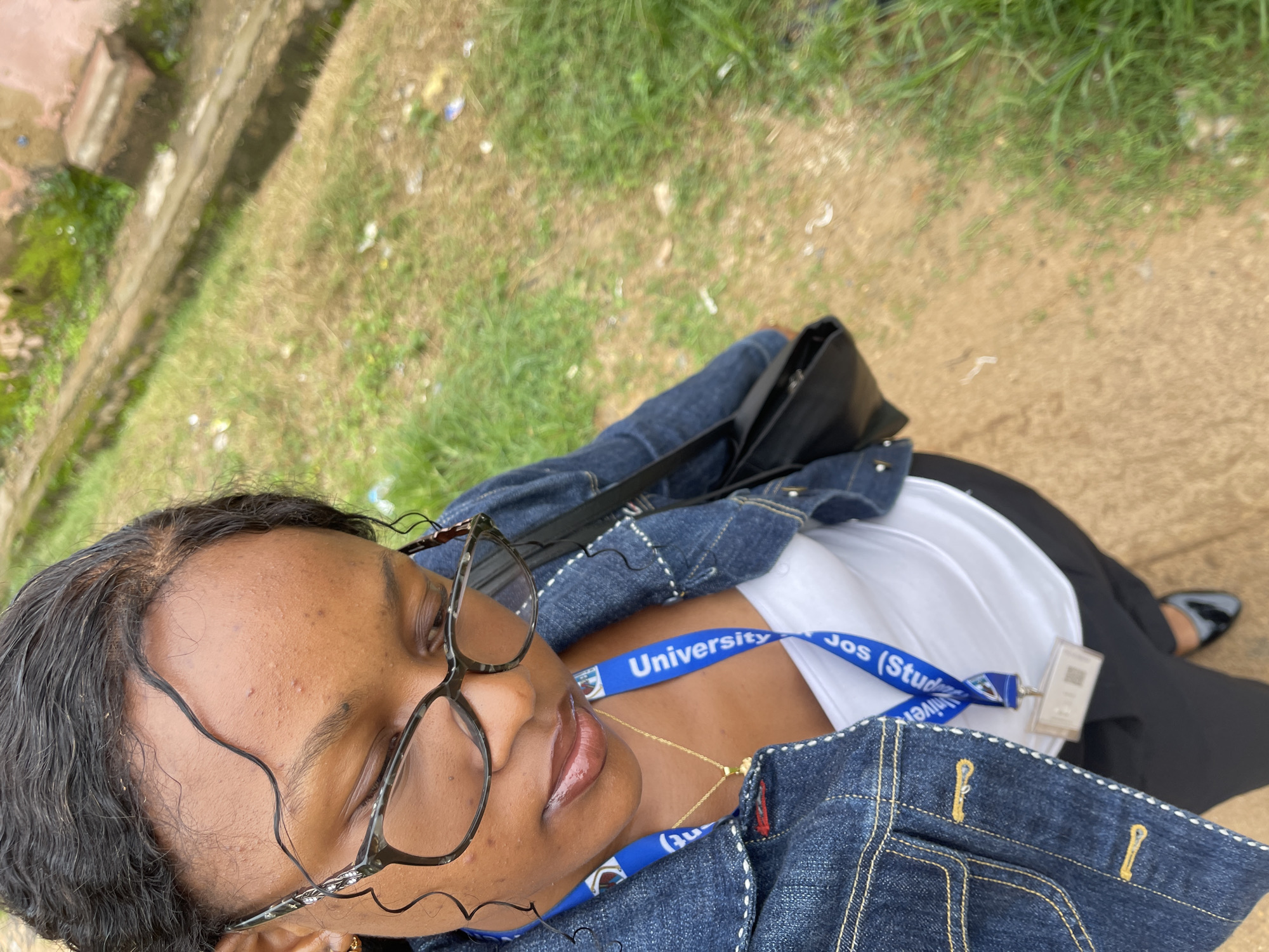 A woman is captured in a close-up, overhead shot taken outdoors in an urban environment. She is wearing glasses, a white top, and a denim jacket. A blue lanyard with "University" visible is around her neck, attached to an ID card. She has a black purse slung over her shoulder. The background consists of a dirt path and overgrown grass, suggesting a less developed area. The image appears to be taken on a cloudy day. The location is identified as Rafin Par, Nigeria.