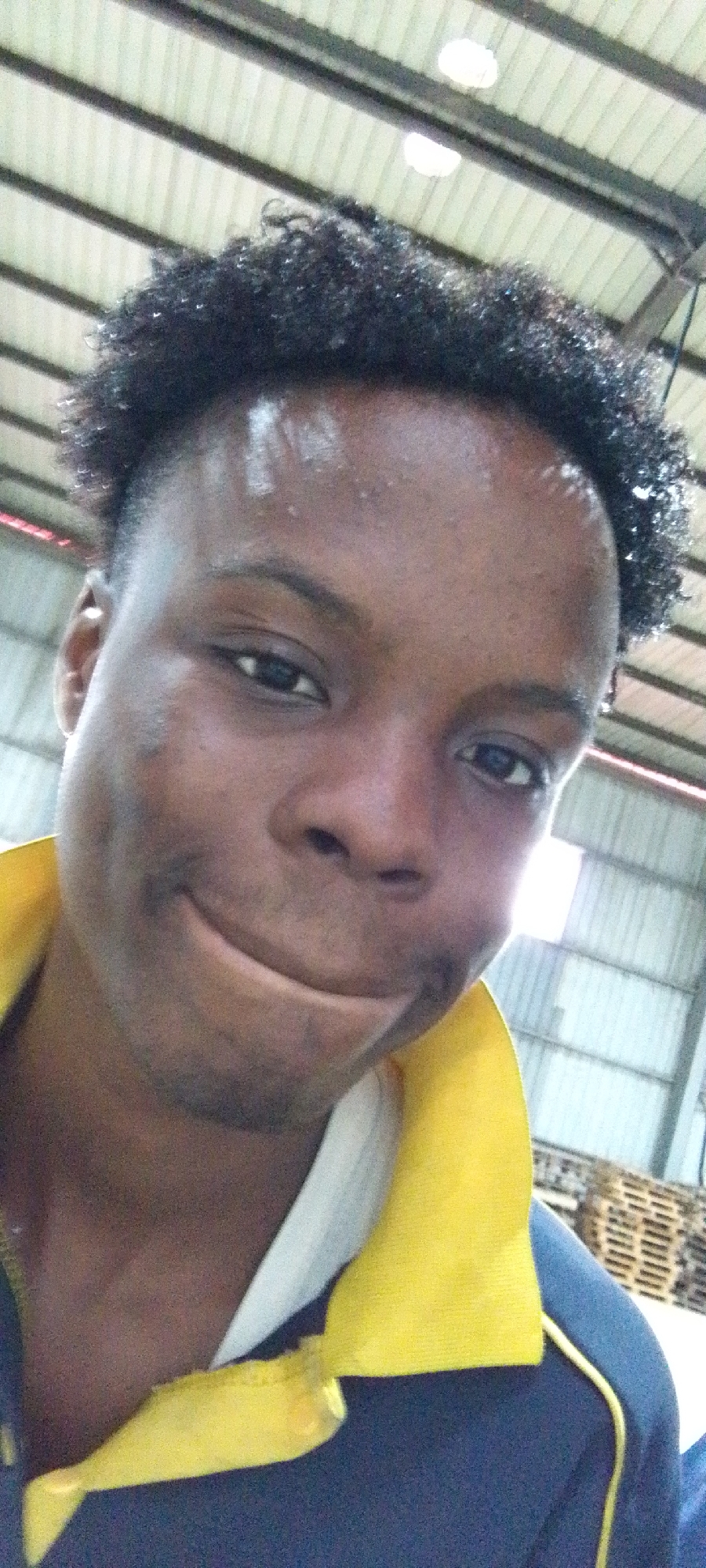 A close-up, slightly upward-angled selfie of a Black male with short, curly, dark hair and a somewhat sweaty forehead. He has dark eyes and is pursing his lips. He is wearing a white undershirt with a dark blue shirt/jacket, possibly with yellow accents or collar.
The background features a corrugated metal roof, suggesting an industrial or storage-type building. Pallets are also visible, indicating storage activity. The location is presumed to be within Ikorodu, Nigeria, based on the prompt.