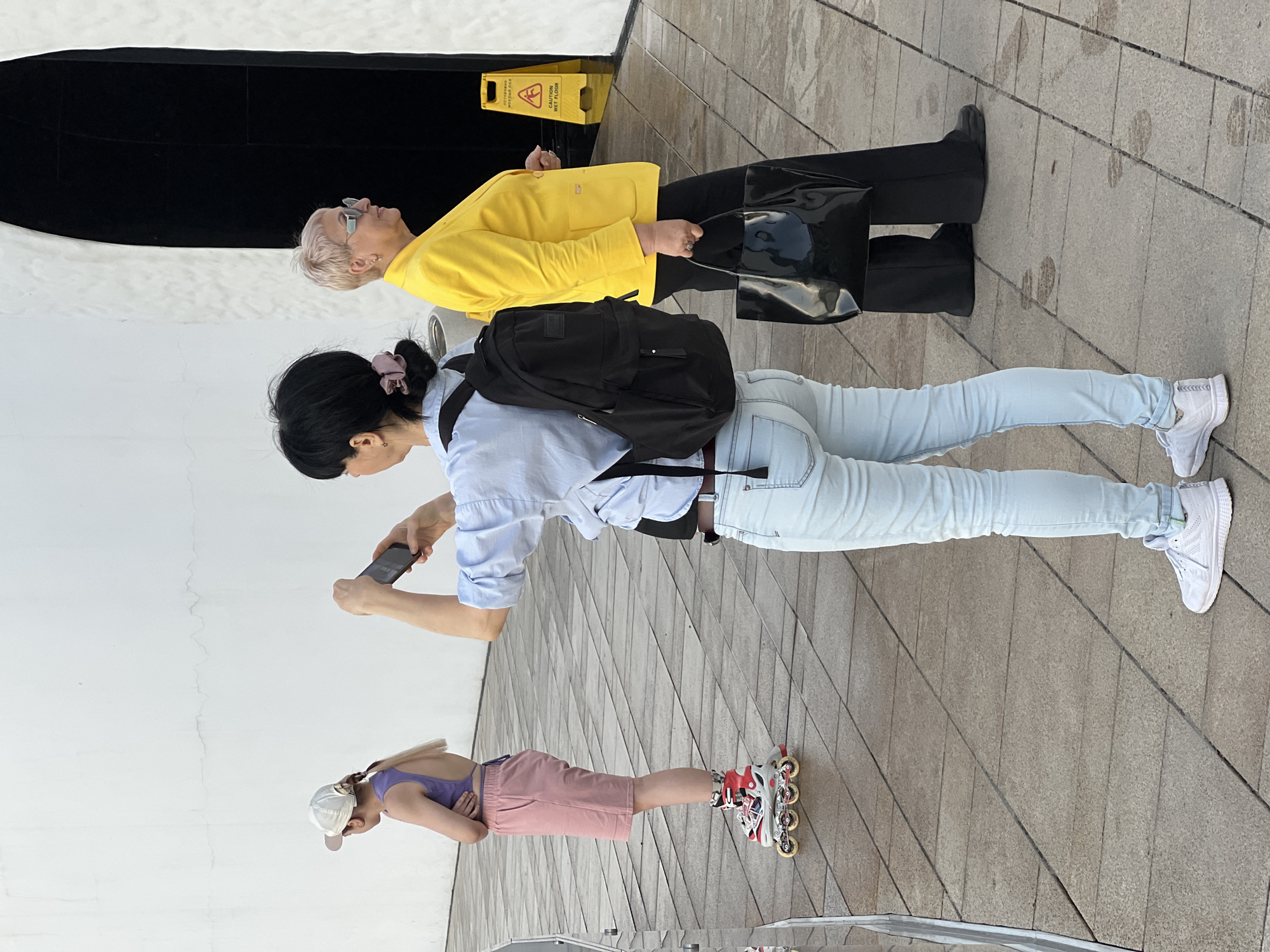 On a paved outdoor area with a herringbone pattern, three people stand near a white wall. The person closest to the wall, viewed from the side, is taking a photo with a smartphone. She has black hair pulled back with a pink hair tie, a light blue button-down shirt, light blue jeans, and white sneakers. She wears a black backpack.
To her right, a woman with short blonde hair wears a yellow blazer, black pants, and black shoes. She carries a black bag. A yellow "Caution Wet Floor" sign sits near the wall.
Further right and in the foreground, a young person on roller skates wears a white baseball cap, a purple top, and a pink skirt.
The scene appears to be in a modern urban setting, possibly Krasnodar, Russia, given the query context. The wall's architecture has a distinctive shape, and the paving is regularly maintained.