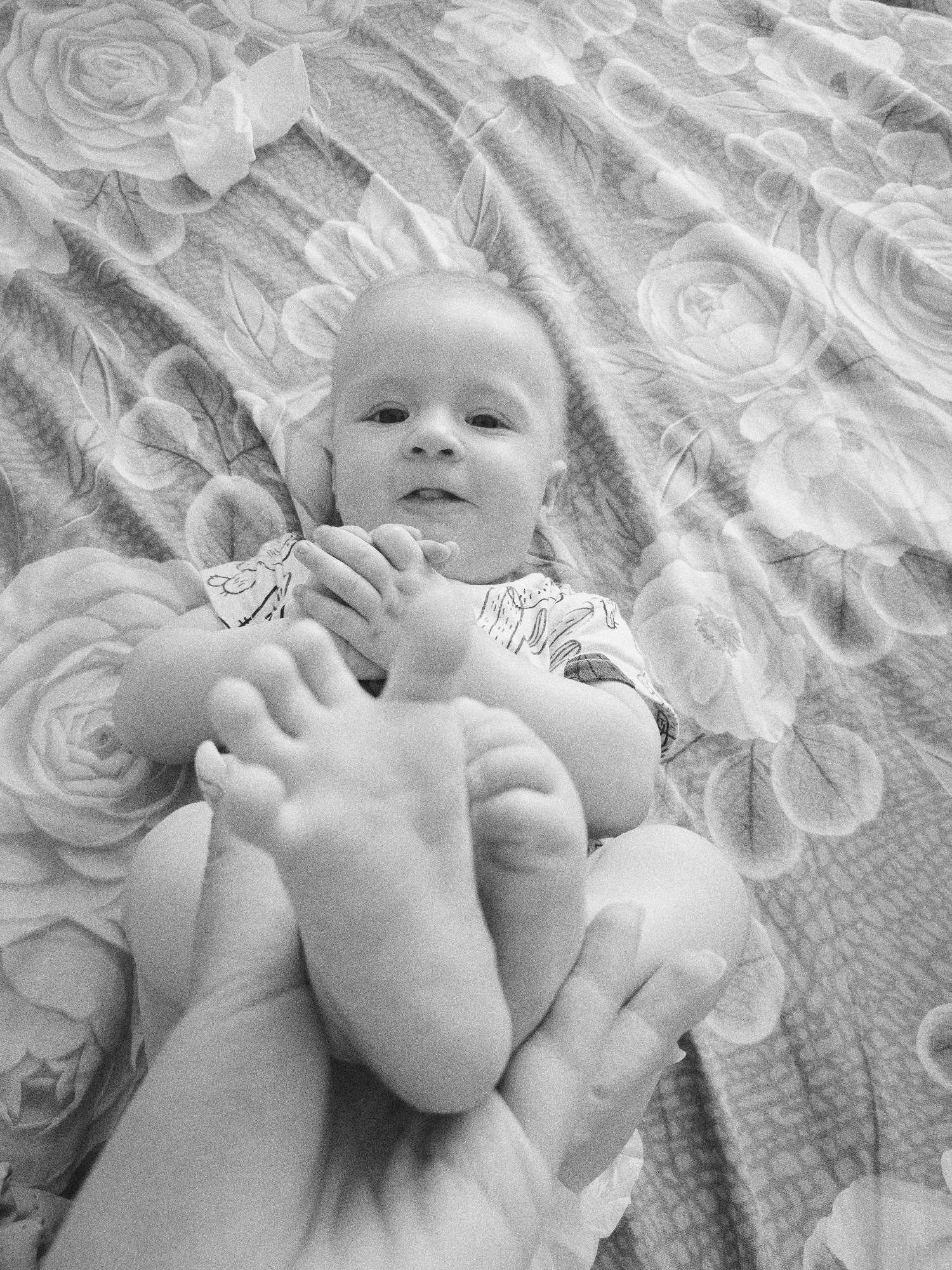 A black and white, medium-shot image depicts a baby lying on a floral patterned surface. The baby, positioned centrally, is on its back and appears to be held by someone. The baby's feet are prominent and held in the hands of an adult. The baby's hands are brought together, close to its face. The baby is wearing clothing. The background appears to be a bedspread or similar textile material, with a pattern of roses. No details suggest this image was taken in Tobolsk, Russia.