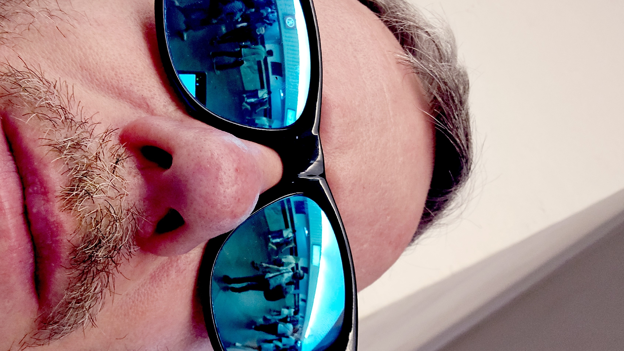 The image shows a close-up of a person's face wearing sunglasses with blue mirrored lenses. The reflection in the sunglasses appears to show a scene of people sitting in chairs, possibly in a lecture hall or waiting room. The individual has gray hair, a mustache, and fair skin. Due to the lack of definitive landmarks and the reflection's ambiguity, the location within Moscow, Russia, cannot be determined with certainty.