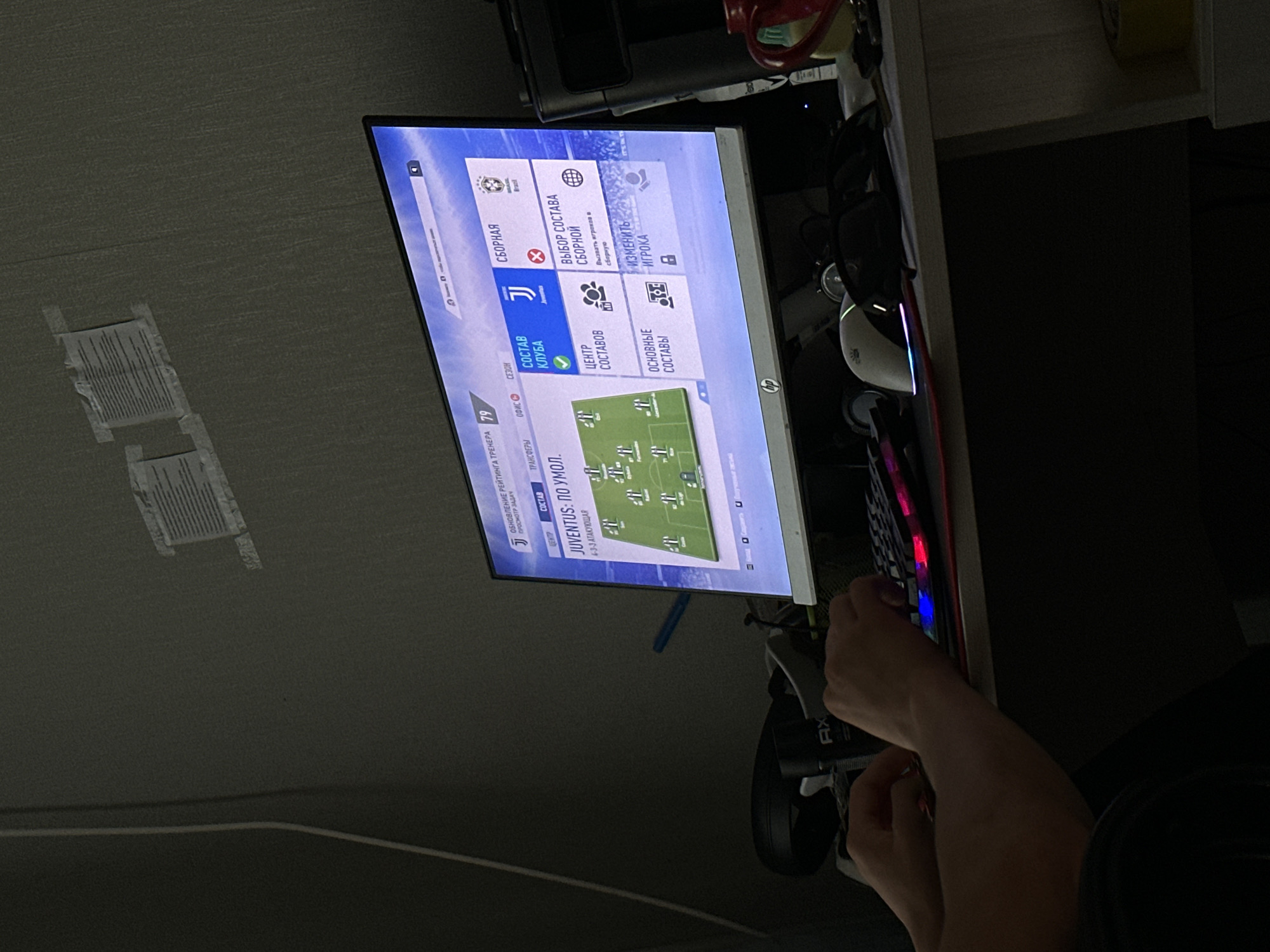 The image shows a person's hands on a keyboard in front of a computer monitor displaying a soccer video game interface, likely FIFA. The monitor shows a team lineup screen for "Juventus" with a 4-3-3 formation. The environment appears to be a dimly lit room, possibly a bedroom or study, with some papers taped to the wall in the background. A desk with a mouse and other gaming accessories is visible. The scene suggests a typical gaming setup. The provided context about the location in Temirtau, Kazakhstan, is not visually verifiable from the image itself.