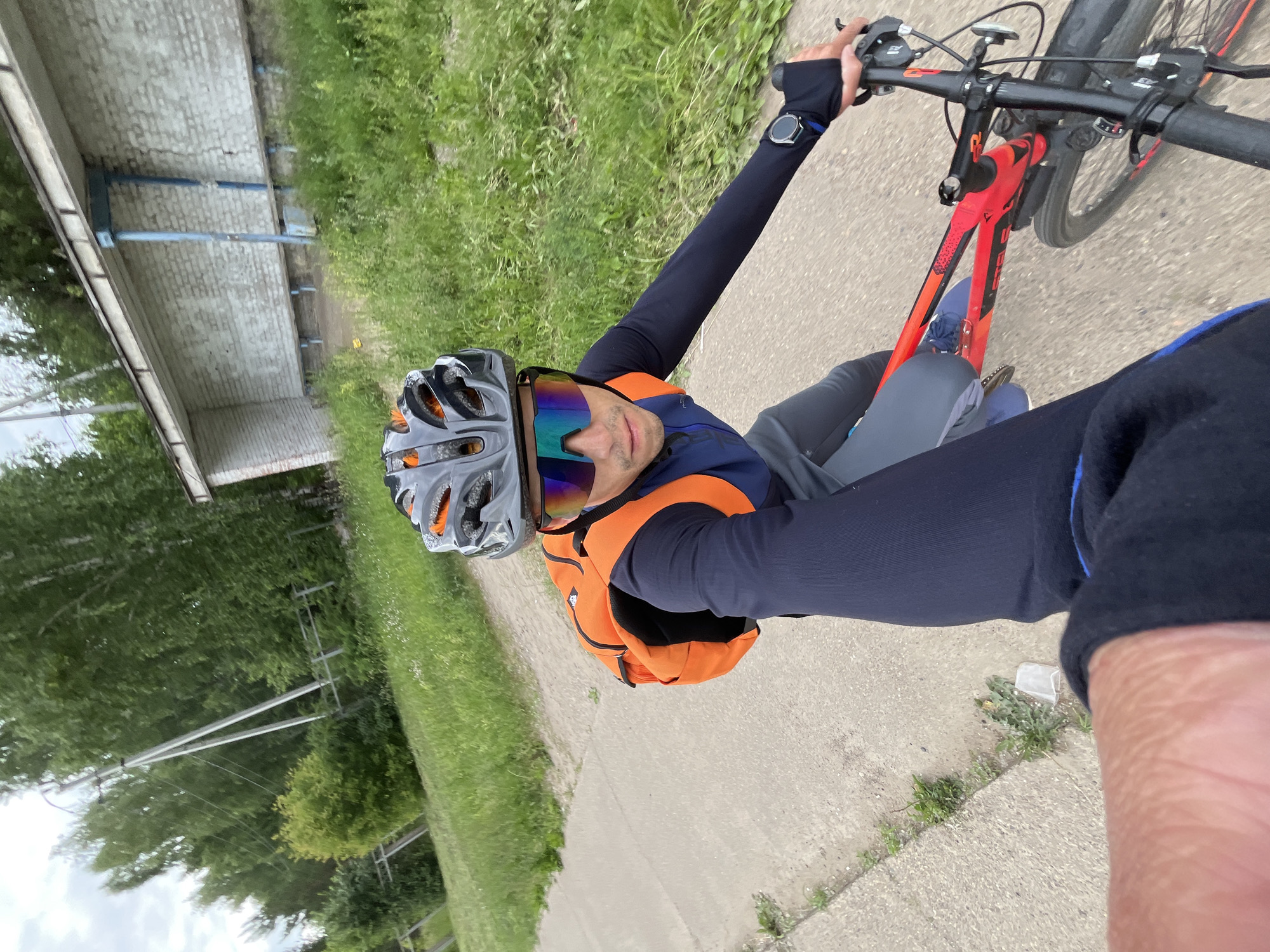 The image shows a first-person perspective of a person riding a bicycle on a paved path. The cyclist is wearing a helmet, sunglasses, a long-sleeved shirt, and pants. They have an orange backpack on. The path is bordered by grass and trees. In the background, there is a brick building with a covered walkway and benches, resembling a bus stop. There are also utility poles and wires visible. The scene appears to be outdoors in a semi-urban or suburban area. The presence of the bus stop suggests a location within a populated area. The architecture of the bus stop, with its white brick and blue supports, is consistent with infrastructure found in many Russian towns and cities. The overall environment suggests a temperate climate with green vegetation.