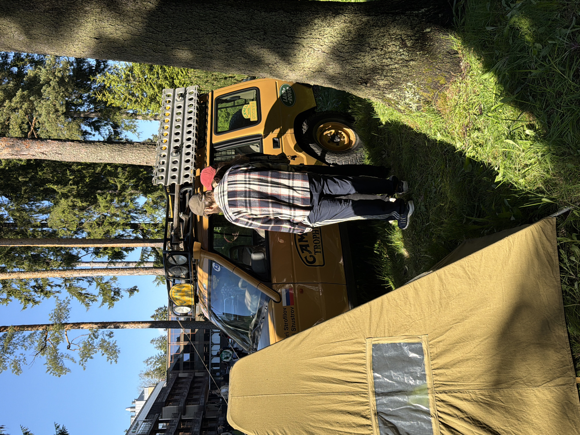 A yellow Land Rover Camel Trophy vehicle is parked next to a khaki-colored tent in a grassy area near pine trees. A person wearing a red hat, a plaid shirt, and Adidas track pants stands next to the vehicle. The vehicle has a rack of metal bars on top and multiple lights. A building is visible in the background. The location is Repino, Russia.