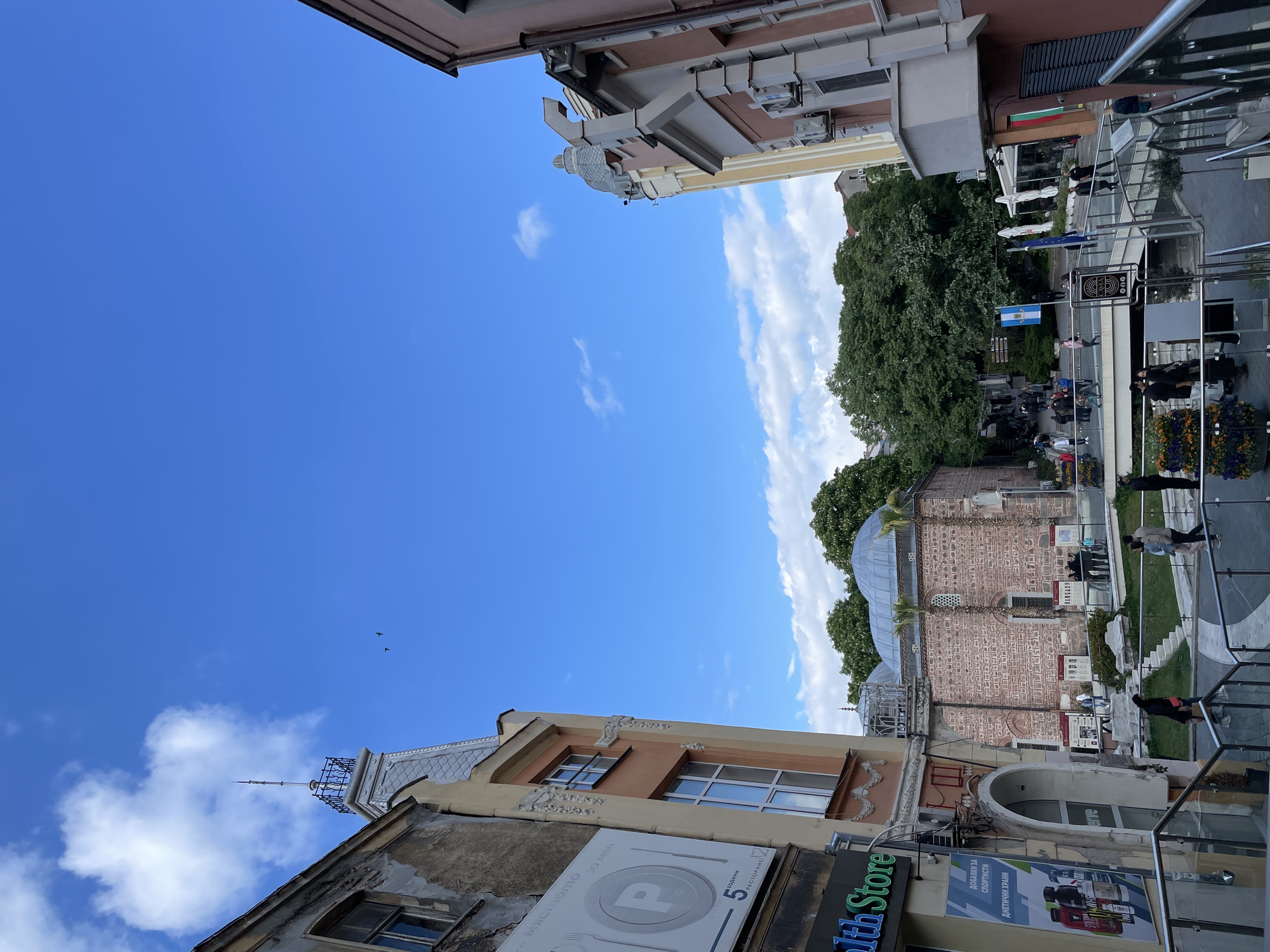 The image shows a street scene in Plovdiv, Bulgaria. Buildings with architectural details are visible on both sides, under a blue sky with scattered clouds. A brick building with a dome, possibly a mosque, is nestled among trees. Pedestrians are present on the sidewalk. Signage for a restaurant ("P") and a health store ("1th Store") can be seen on one of the buildings. Flags are visible in the upper right, including the Bulgarian flag. The scene includes a mix of old architecture and modern elements, such as glass railings.