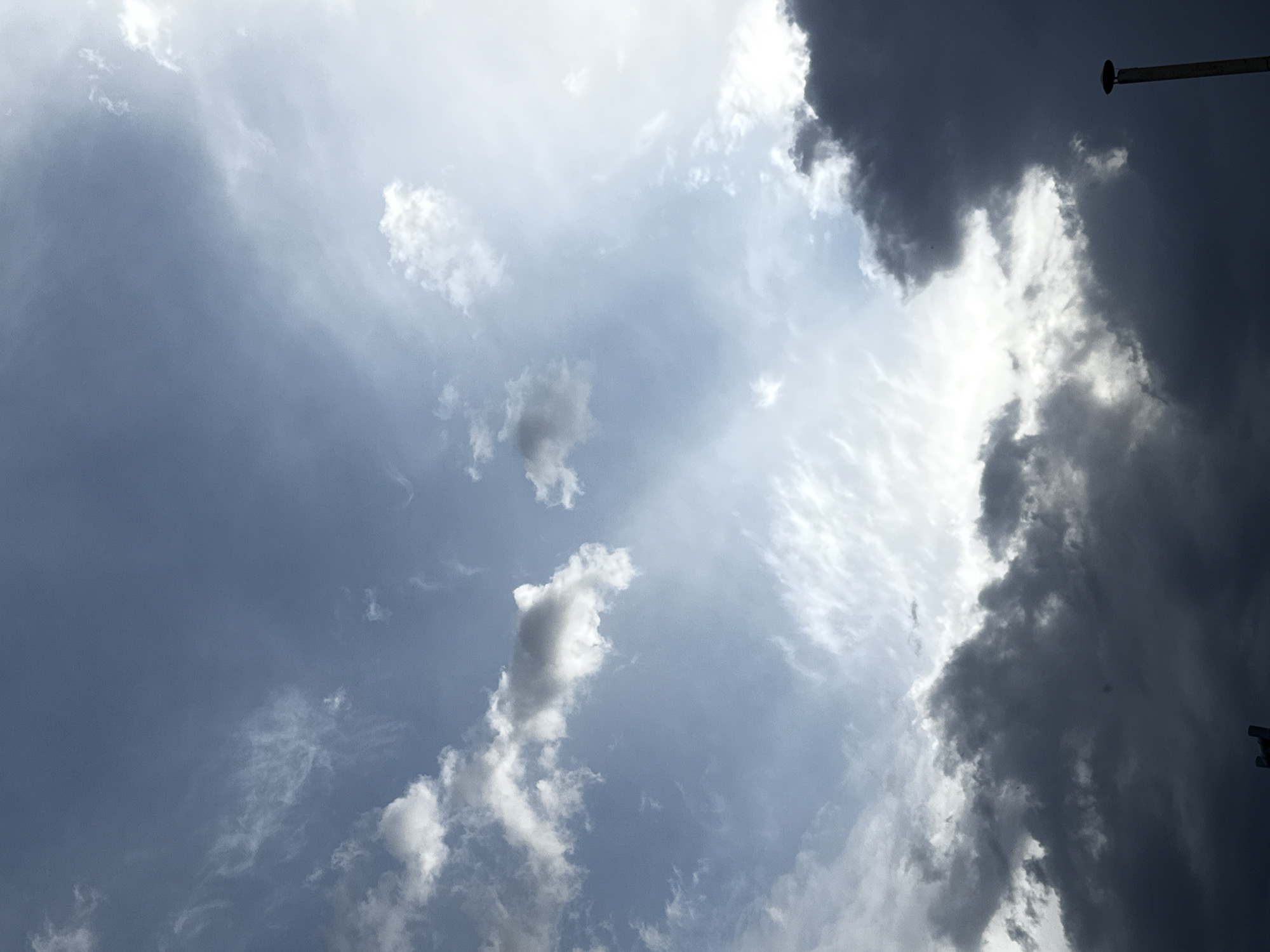 The image depicts a sky with a mix of light and dark clouds. A section of the sky appears overcast with dark, ominous clouds massing on the right side of the frame. Conversely, the center and upper left of the image reveal lighter, brighter sky with scattered white clouds. Part of a tall pole is also present in the top right corner. The location is unknown.
