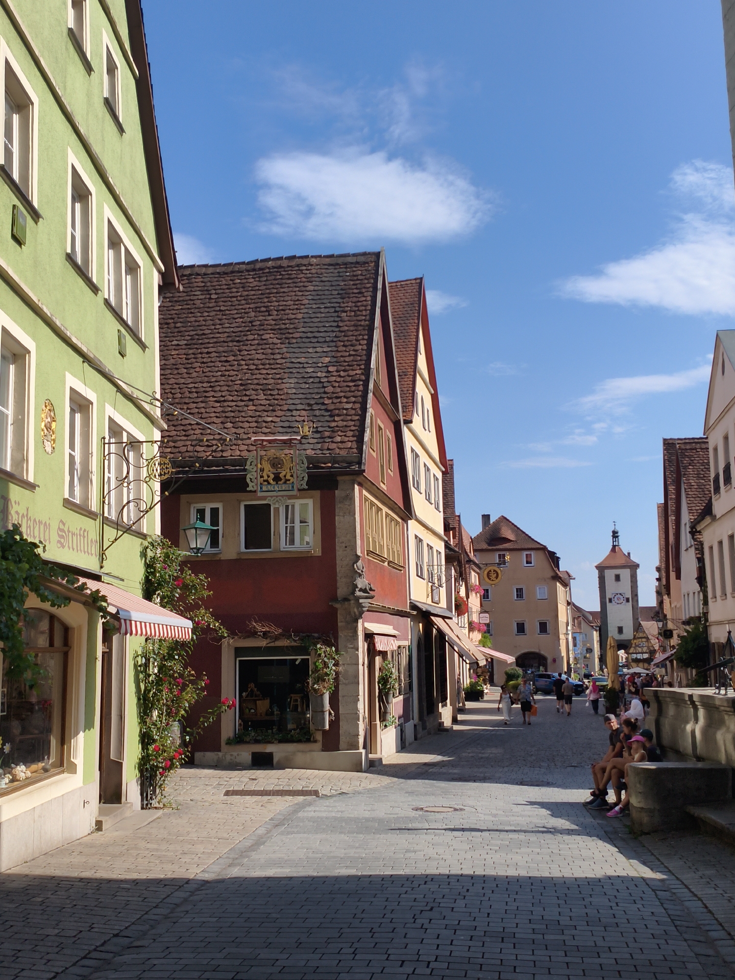 The image depicts a view of a cobblestone street in Rothenburg ob der Tauber, Germany, likely in the heart of the historic old town. On the left, a light green building with cream-colored trim and numerous windows lines the street. A sign with German text is partially visible on this building, along with a decorative wrought-iron bracket. Red and white striped awnings are present above some windows.
To the right of the green building, a series of other historic buildings with steeply pitched, red-tiled roofs and colorful facades (red, yellow, and pale orange) are visible. These buildings feature arched doorways and windows, characteristic of medieval architecture. Some of these buildings appear to be shops, with display windows and awnings.
Further down the street, a distinct tower with a clock face, a common feature in German medieval towns, stands in the distance. People are seen walking and sitting on a stone bench along the right side of the street, enjoying the sunny weather. The sky is blue with scattered white clouds. The overall scene conveys a vibrant and picturesque atmosphere of a well-preserved historic European town.