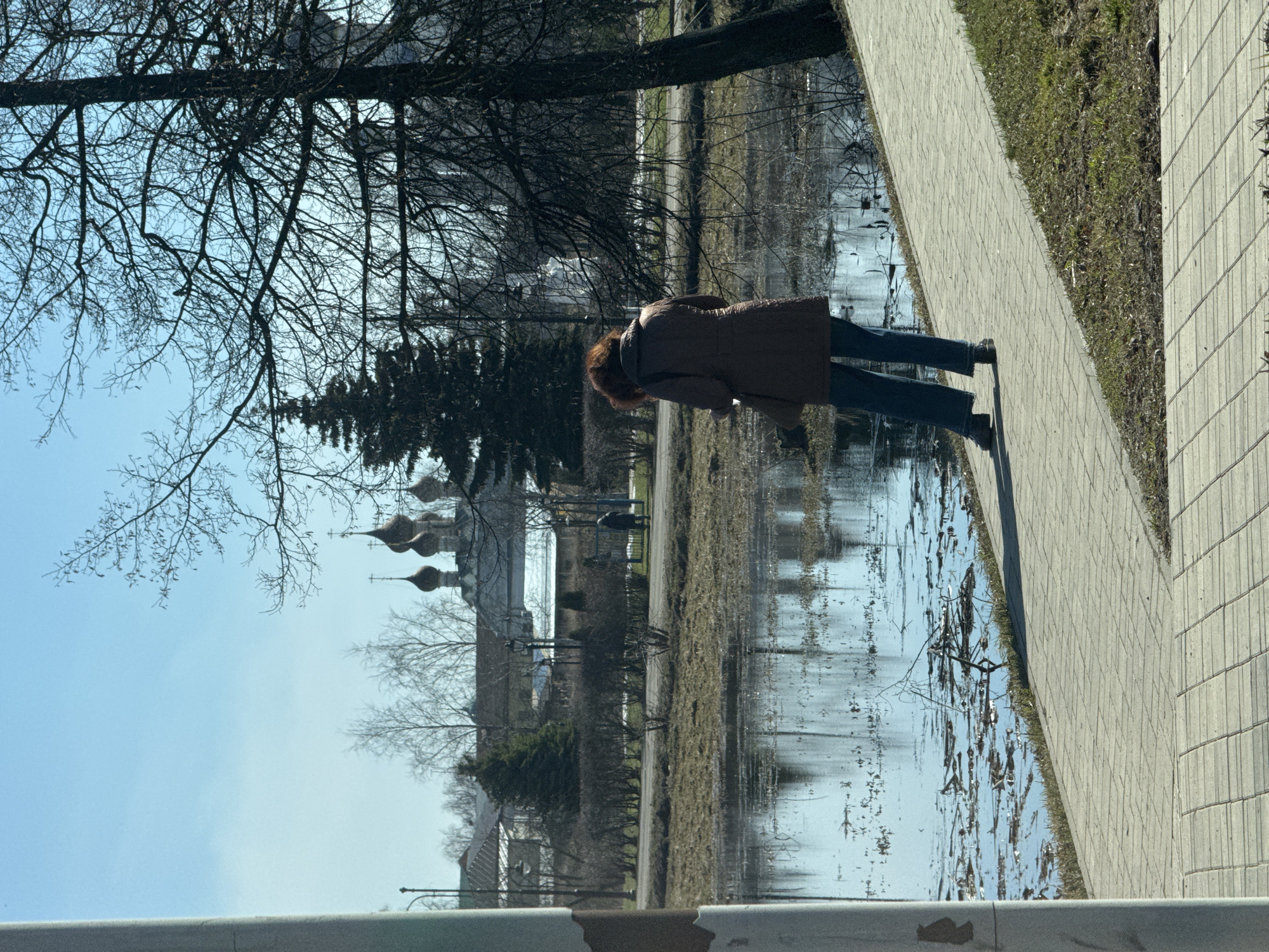 A woman in a brown coat and blue jeans walks along a brick sidewalk next to a body of water, possibly a flooded area or canal, with vegetation visible along the edge. The reflection of bare trees and buildings can be seen in the water. In the background, there are structures with distinctive domes, indicating a Russian Orthodox church, and trees with bare branches. The sky is blue. The scene appears to be in a urban park setting. The image captures a moment of everyday life in an area with prominent architectural and natural features. The image is taken in Ново-Никольское, Russia.