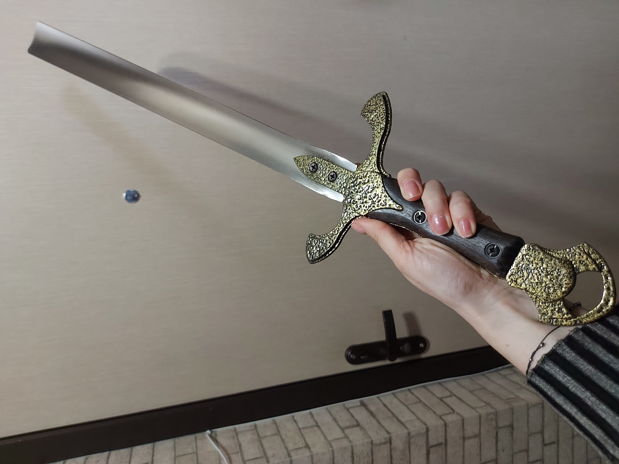 A hand is holding a decorative sword. The sword has a silver-colored blade, a dark brown handle, and a gold-colored guard and pommel with ornate detailing. The hand has pale skin and manicured nails. The background includes a light-colored door with a dark brown trim, a door handle, and a tiled surface along the bottom. The scene is not identifiable as specifically located in Izhevsk, Russia or any other specific location.