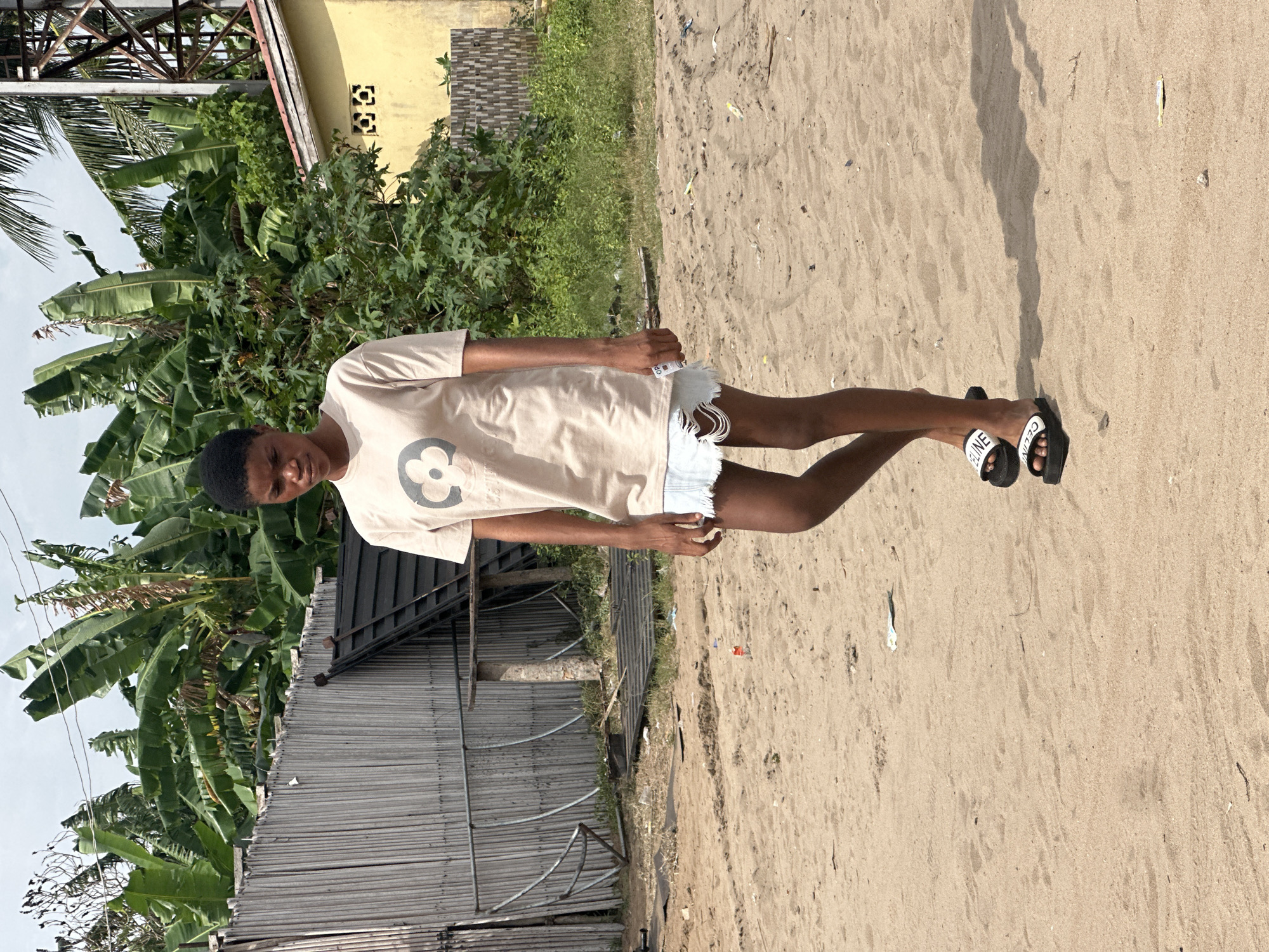 A young person with dark skin and short dark hair stands facing the camera, slightly smiling. They are wearing a light beige t-shirt with a dark, circular logo (resembling a stylized 'C' or 'G') on the chest, and light-colored denim shorts with frayed edges. On their feet are black slide sandals with "CELINE" written in white across the straps. The person's right arm is bent, holding a small white object with "OP" visible on it.
The setting is outdoors, characterized by sandy ground covering the foreground and midground. Behind the person, there's lush green vegetation, including large banana leaves, palm fronds, and other leafy plants. In the background, man-made structures are visible, including a grey corrugated metal fence or wall, parts of a yellow building with square decorative vents, and a metal framework or structure. The sky is bright, suggesting clear weather and daylight, with distinct shadows cast on the sand by the person. The overall scene suggests a warm, possibly tropical, outdoor environment, consistent with a location like Badagry, Nigeria.