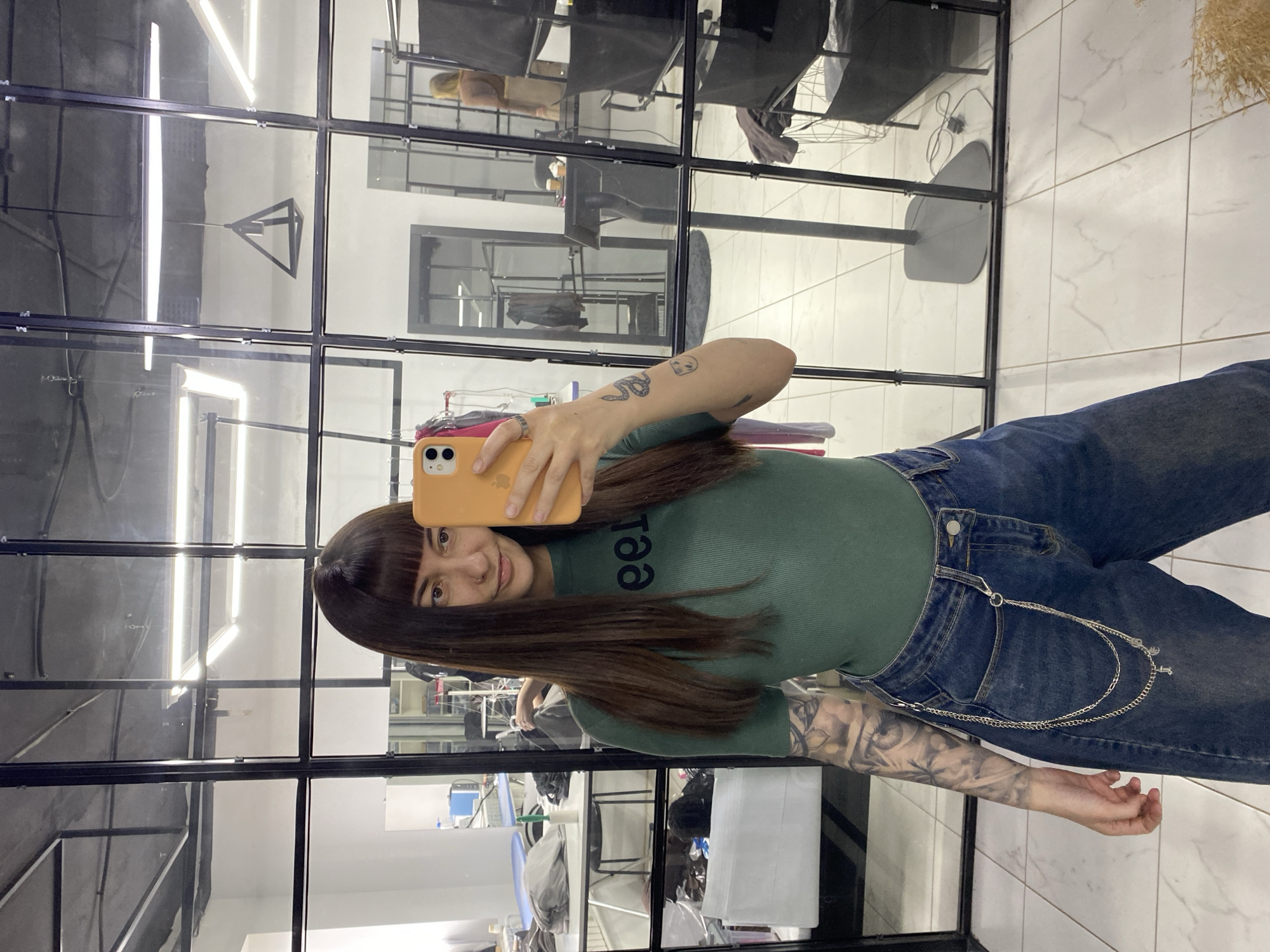 A young woman is taking a selfie in what appears to be a hair salon or a beauty parlor. The woman has long brown hair with bangs, is wearing a green top, and blue jeans with a chain detail. Her left arm is visible and has tattoos. She is holding an orange smartphone to take the picture. The setting is a commercial space with a tiled floor and a series of black-framed glass panels forming a wall or partition. The glass panels reflect the interior of the salon, showing chairs, lighting fixtures, and other salon equipment. Given the prompt instructions for location, the image is presumed to be in the city of Votkinsk, Russia.