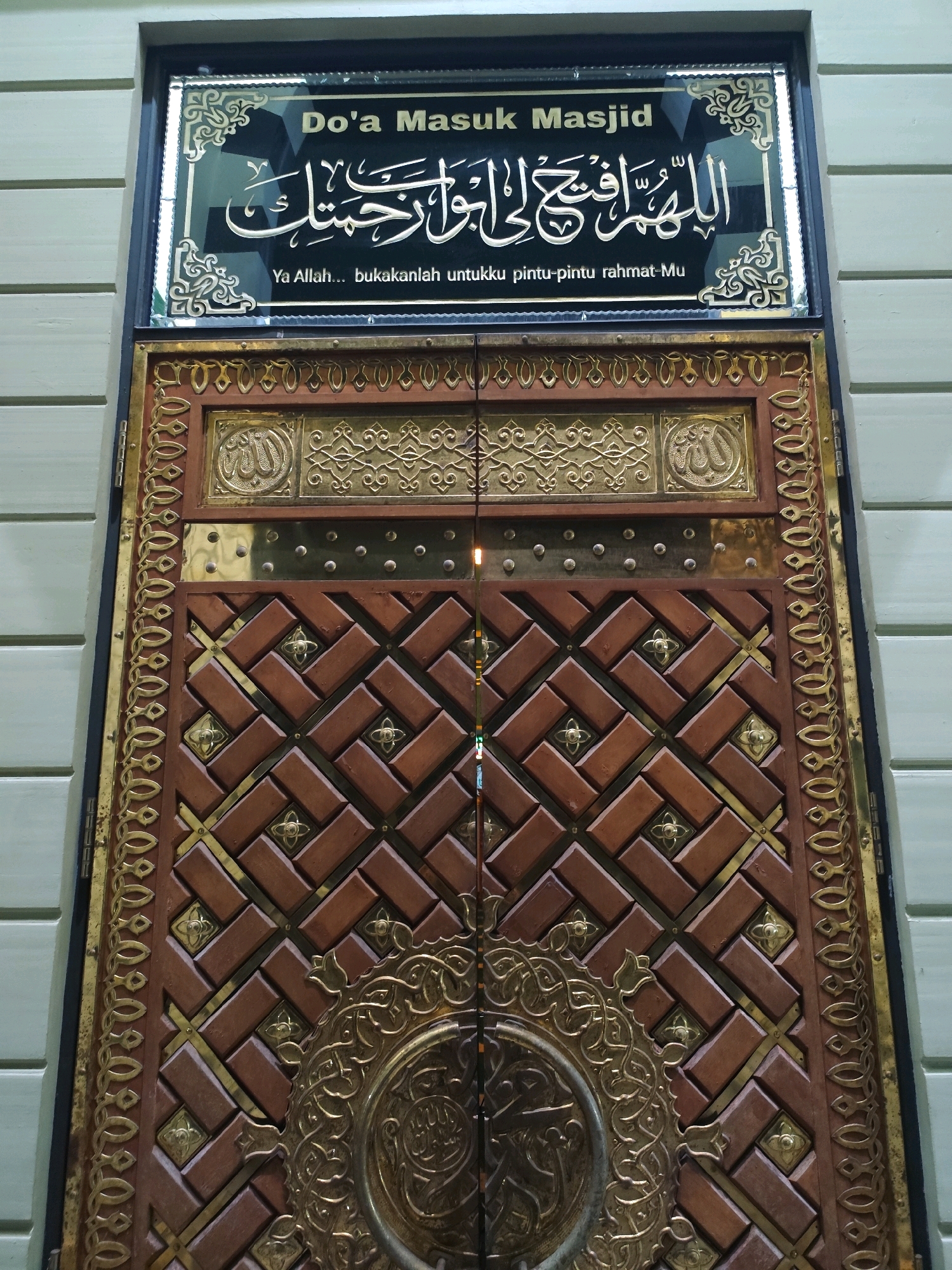 A close-up, slightly angled shot shows the ornate entrance to the Masjid Jami' Palangka Raya in Palangka Raya, Indonesia. The upper part of the image features a black rectangular plaque with gold Arabic calligraphy and text in English. The Arabic text translates to "O Allah, open for me the doors of Your mercy." Below this, in English, it reads "Ya Allah... bukakanlah untukku pintu-pintu rahmat-Mu" which also translates to "O Allah... open for me the doors of Your mercy." The dominant feature of the image is a large, intricately carved wooden door with brass accents and decorative patterns. The door is divided into two panels, each adorned with a repeating geometric motif of interlocking brown wooden blocks and smaller golden floral accents. A prominent circular brass emblem is situated at the center of the lower portion of the door. The surrounding walls of the building are visible on the left and right, featuring a light green horizontal paneling. The lighting suggests it is daytime.