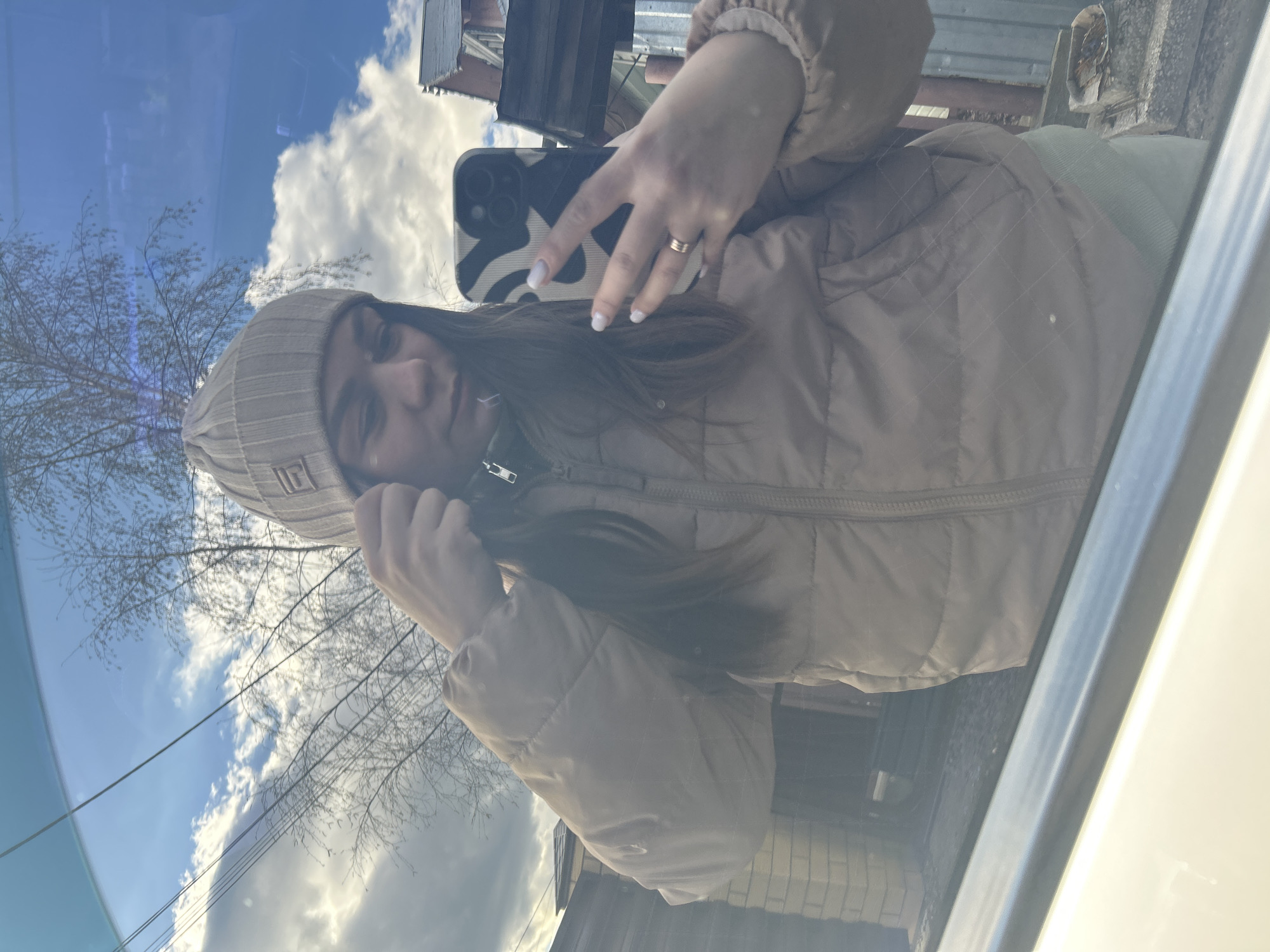 The image captures a young woman taking a selfie, reflected in what appears to be a window or glass surface, likely from inside a vehicle. She is seen from the chest up, with long, dark hair, wearing a light beige or tan puffer jacket and a matching ribbed beanie hat. A small, indiscernible logo or text is present on the front of her beanie.
She is holding a smartphone in her right hand to take the picture. Her hand shows light-colored nail polish and a simple gold-toned ring on her ring finger. The smartphone case features a distinctive black and white abstract or cow-print design.
The reflected background reveals a bright, clear blue sky with white, fluffy clouds. Bare tree branches, suggesting a late autumn, winter, or early spring season, are prominent in the reflection, along with power lines. Glimpses of distant buildings, possibly residential or small structures, are also visible.
The scene is set during the daytime under sunny weather conditions. Parts of the vehicle's interior are visible in the foreground and along the right edge, including what looks like a seat or dashboard area, and another jacket, possibly dark green or gray, on the seat beside her. The woman's expression is relaxed. The location context is Chaikovsky, Russia.