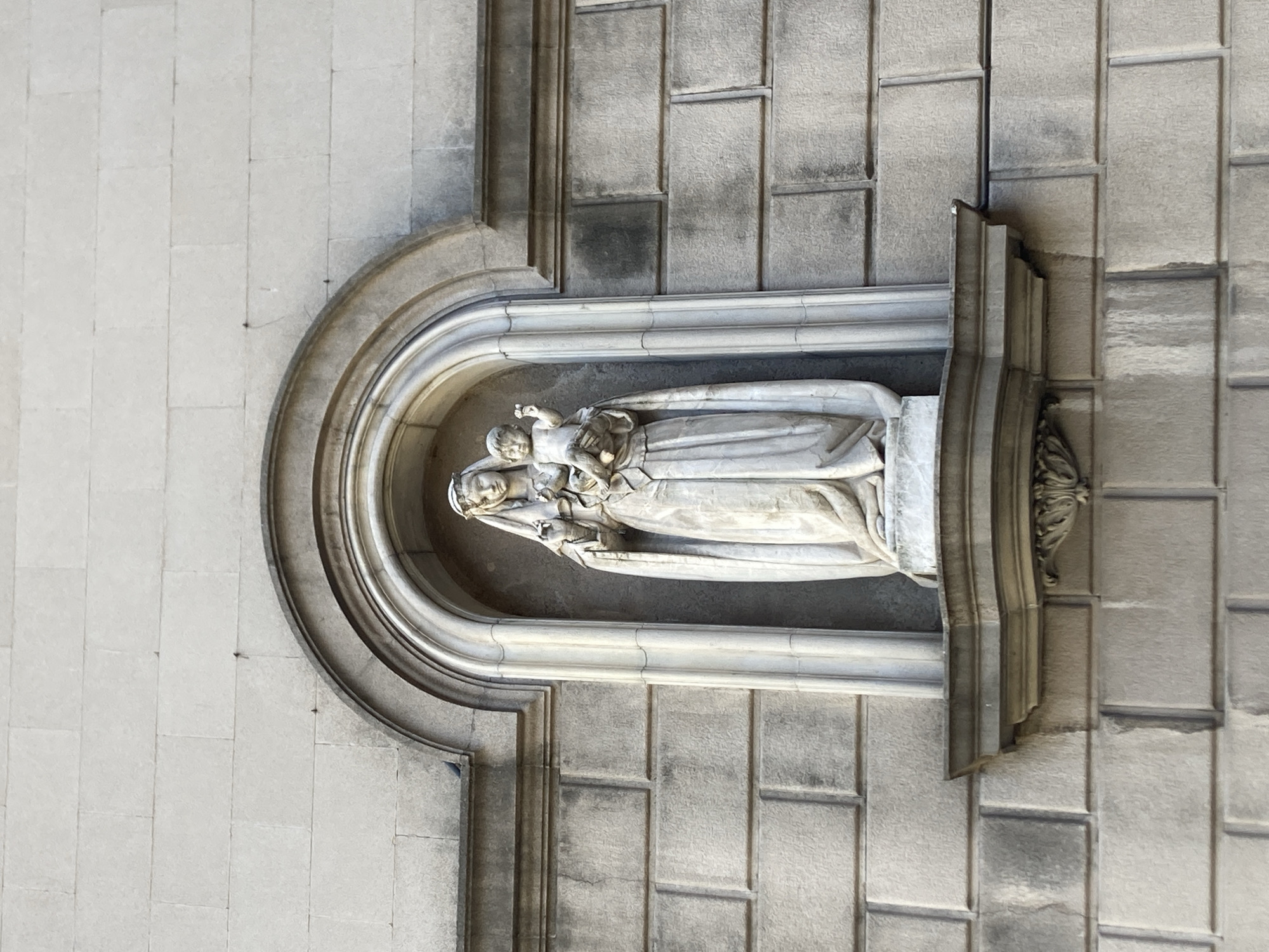 This image captures a stone statue of a woman and child, carved in a classical style. The figures are set within a semicircular niche that is recessed into a stone wall. The woman, depicted as a serene Madonna, cradles the infant Jesus in her arms. Both figures are draped in flowing robes, characteristic of religious iconography. The statue appears aged, with some wear and tear visible, suggesting it has been exposed to the elements for a considerable time.
The surrounding wall is constructed from large, rectangular stone blocks, laid in a grid pattern. The niche itself is framed by a more elaborate, decorative molding, also made of stone, with curved architectural elements. To the right of the niche, a section of the wall features more ornate stonework, including what appears to be a decorative carving or corbel.
The lighting suggests it is daytime, with sunlight casting shadows that highlight the texture and depth of the stone carvings and the wall. The overall scene conveys a sense of timelessness and reverence, typical of religious art and architecture. Given the prompt's location context of Barcelona, Spain, this statue is likely part of a historical building, possibly a church or a public monument in the city. No visible text is present in the image.