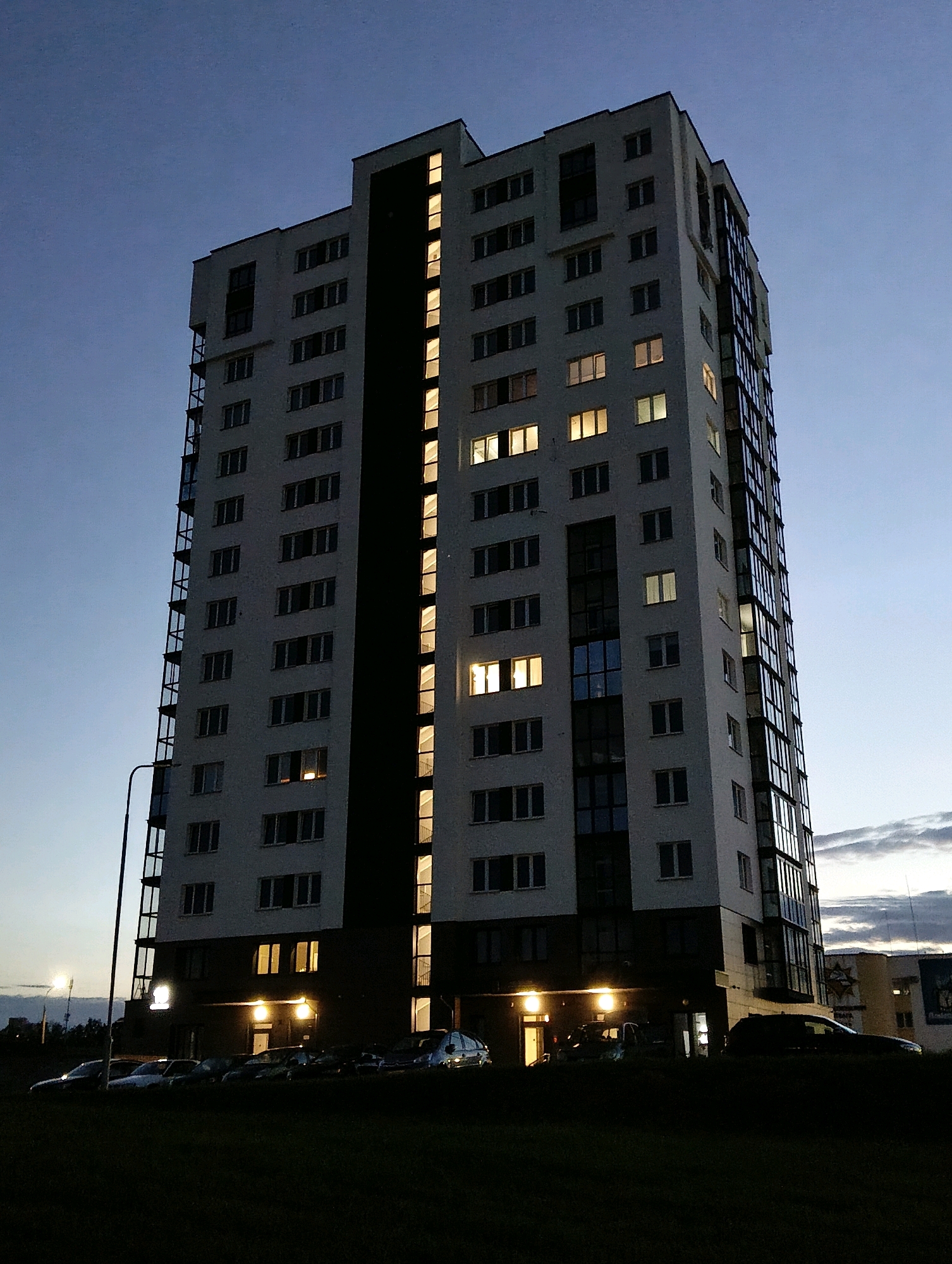 The image shows a modern, multi-story apartment building in the city of Hrodna, Belarus, during twilight. The building is predominantly white with dark vertical sections and a glass facade on one side. Numerous windows are visible, with several illuminated from within, indicating occupancy. At the base of the building, a covered entryway is lit, and several cars are parked in the foreground. The surrounding area appears to be a grassy field with a street lamp visible to the left. The sky is a deep blue, transitioning to lighter hues near the horizon, with some scattered clouds.