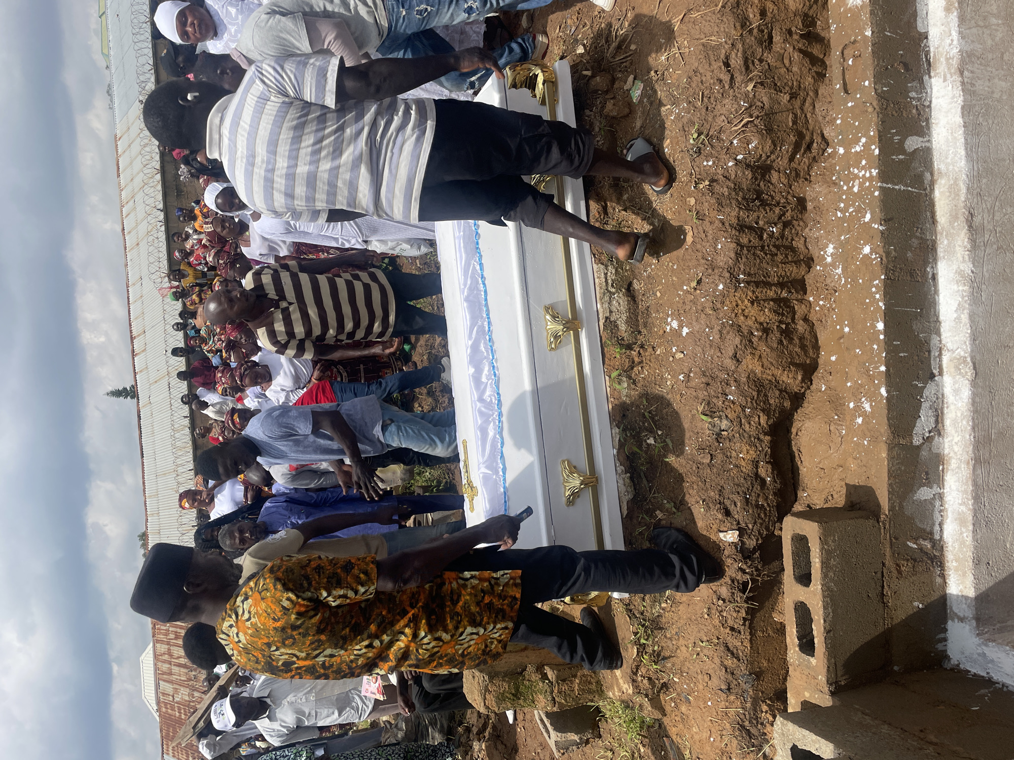 In Gura Top, Nigeria, a funeral procession is underway. A white coffin with gold accents rests in a freshly dug grave. The coffin is surrounded by a gathering of mourners, some of whom appear to be actively involved in the burial. The environment is outdoors, with evidence of an ongoing construction or burial site. The sky is cloudy, suggesting potentially overcast weather. The scene is somber, reflecting the nature of a funeral ceremony.