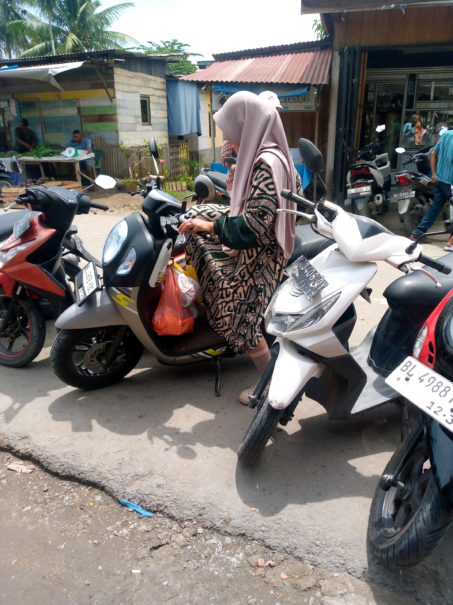 In Keudeteupinpunti, Indonesia, a woman wearing a light pink hijab and a patterned dress is standing next to a grey scooter. The scooter has a red plastic bag hanging from its handlebars, containing orange items. To the left of the grey scooter is a red scooter. In the background, there is a small building with a corrugated metal roof and wooden walls, appearing to be a roadside stall or shop. Several other scooters are parked behind the main subjects, and a few people are visible further in the background, some appearing to be customers or vendors. The scene is set outdoors on a paved surface with some debris on the ground, under a bright sky with palm trees visible.