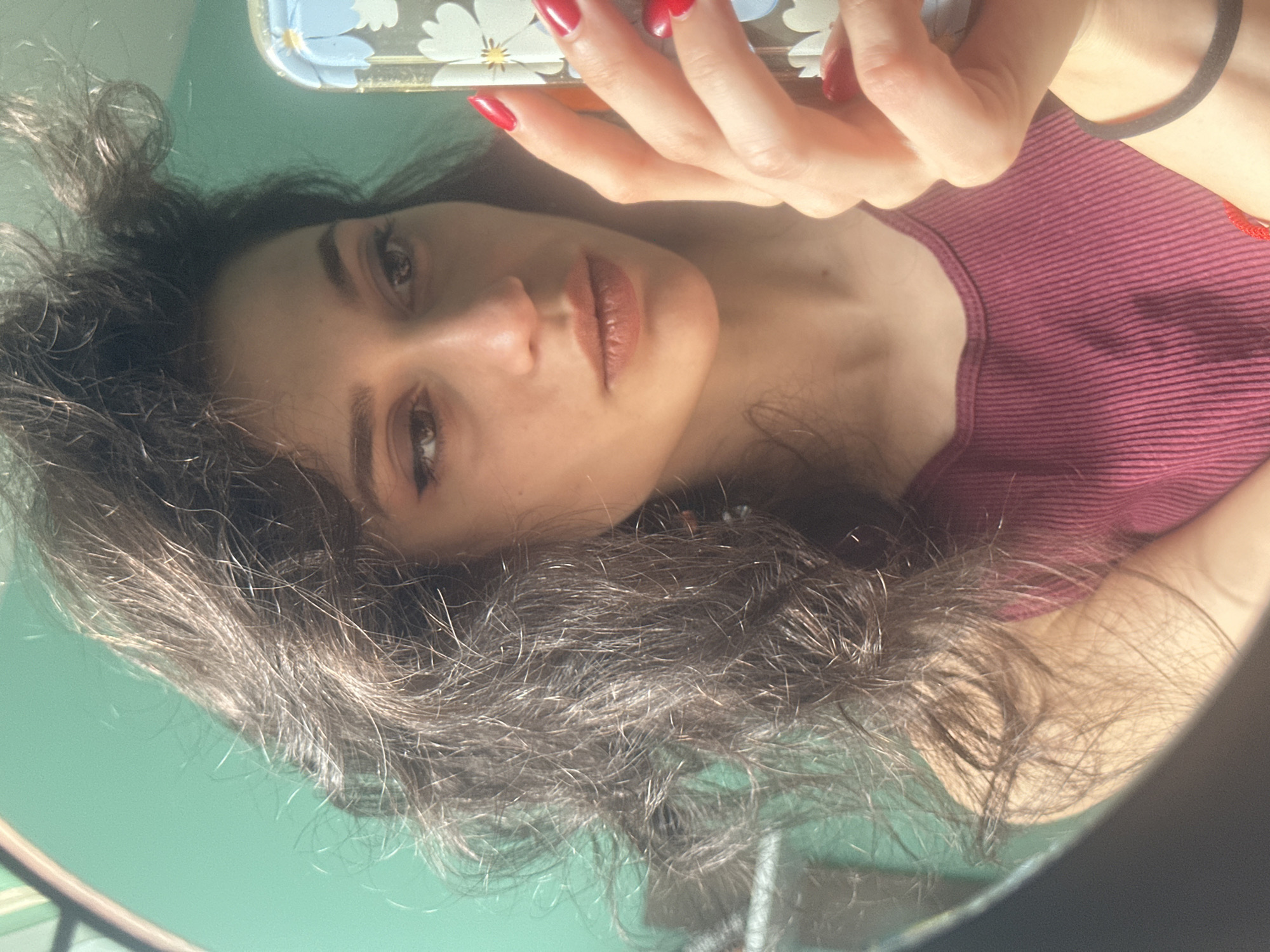 A young woman with dark, wavy hair, wearing a maroon ribbed tank top, takes a selfie in a mirror. Her right hand, with bright red nails, holds a smartphone with a floral pattern. She has on subtle makeup, including winged eyeliner. The background is a solid teal wall, illuminated by natural light casting shadows on her hair. The image is a close-up, focusing on her face and upper body. The reflection shows the side of her head and a hint of her shoulder. The context of this image being taken in Sofia, Bulgaria, is not discernible from the visual information provided in the image.