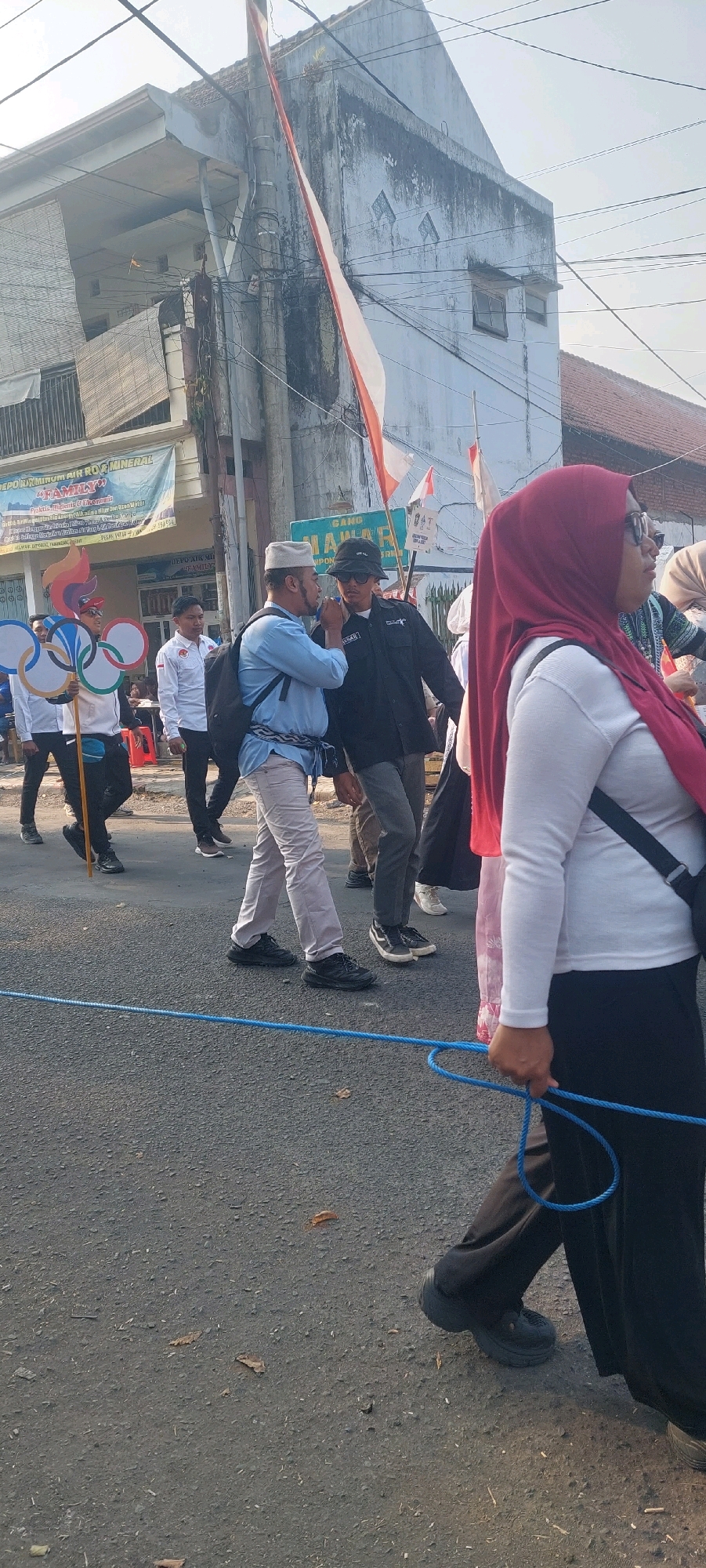 In Jogoyudan, Indonesia, a procession is underway on a paved street. A person on the left holds a large cutout of the Olympic rings. Several other individuals are walking in the procession, including a man wearing a light blue shirt and a white skullcap, who is speaking to another man wearing a black jacket and a dark hat. A woman in the foreground wears a red hijab, a white long-sleeved shirt, and black pants. Other participants are visible in the background, as are buildings and flags. The environment suggests an outdoor event on a sunny day.