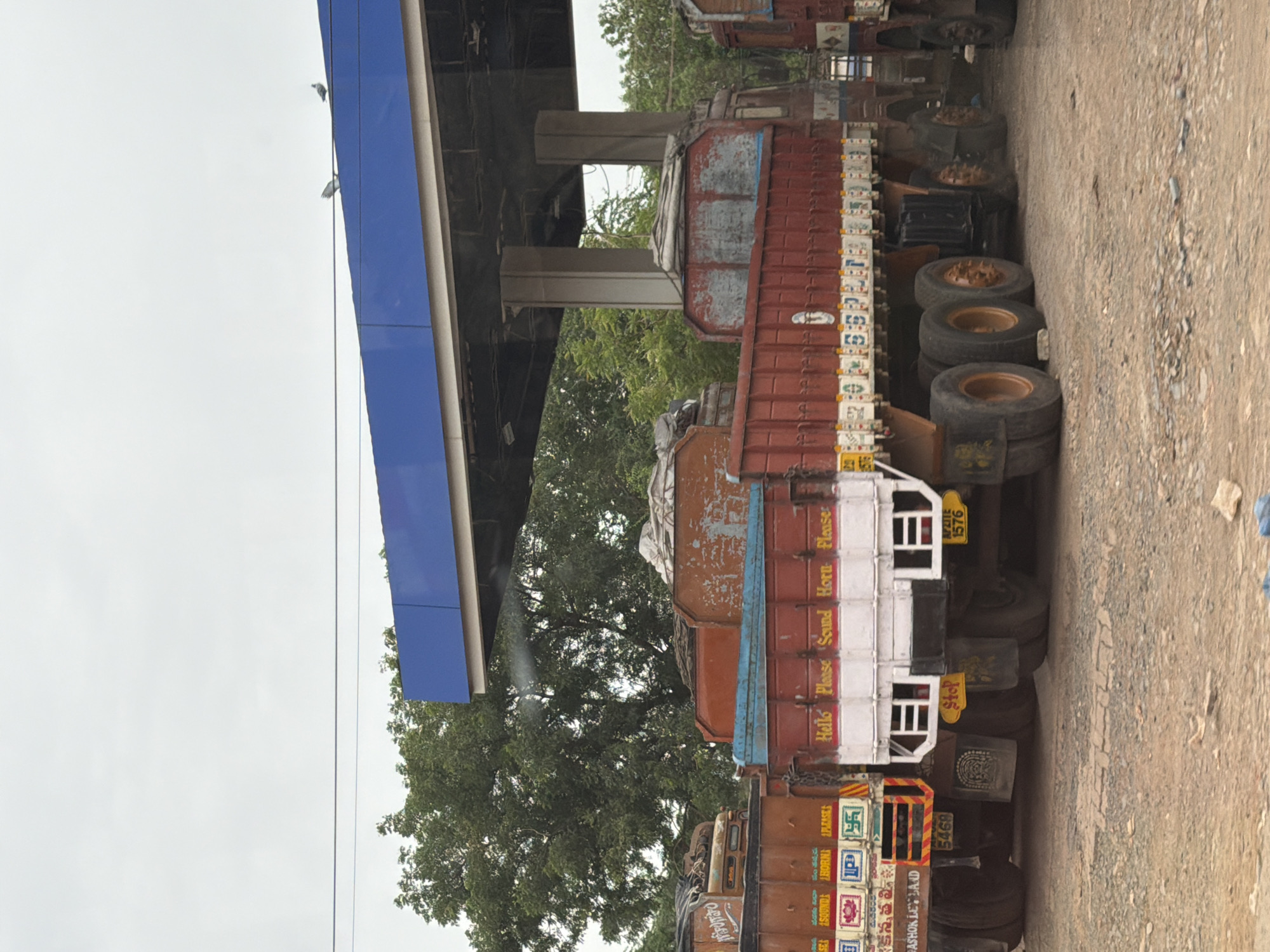The image depicts a row of parked trucks under a blue canopy structure, likely a covered parking area or a fuel station, in Bethamcherla, India. The trucks are predominantly red and brown, with visible wear and tear. Some trucks have white lettering on their sides, including the phrases "Hello Please Sound Horn Please". The ground is a mixture of dirt and gravel. In the background, lush green trees are visible, suggesting an outdoor setting. The sky is overcast. Two pigeons are perched on the blue canopy. The trucks appear to be commercial vehicles, possibly for transport or logistics. No people are visible in the image.