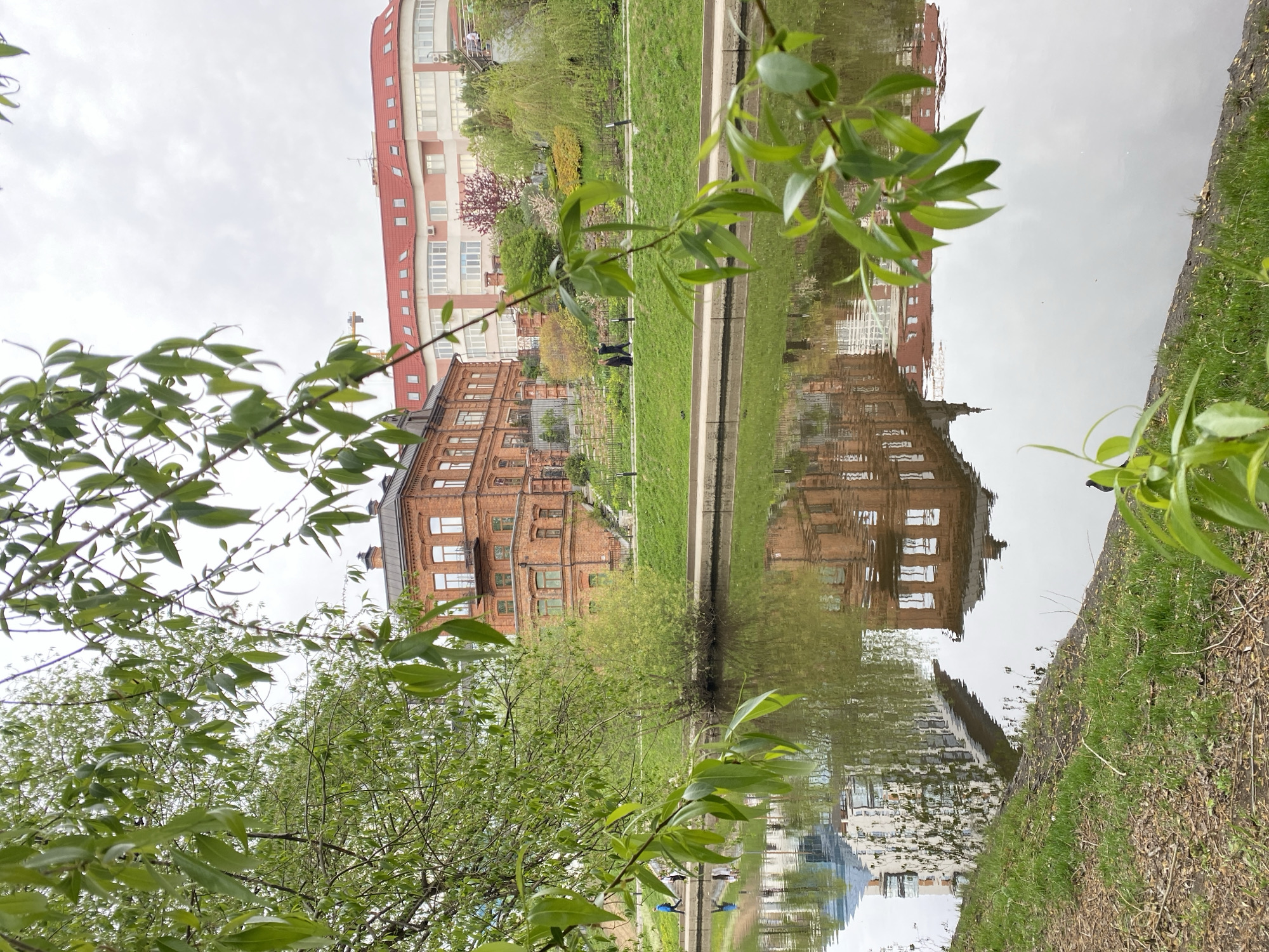 A ground-level, rotated landscape photograph depicts a scene in Yekaterinburg, Russia, featuring a body of water reflecting buildings. The buildings include a red brick building with a dark roof and multiple windows, and a modern building with a red roof and white facade. A narrow strip of green grass separates the buildings from the water's edge. The foreground is dominated by tree branches with green leaves, obscuring parts of the scene. The water's surface reflects the buildings, creating a mirror image. The sky is overcast, suggesting a cloudy day. Along the edge of the water is dirt and grass.