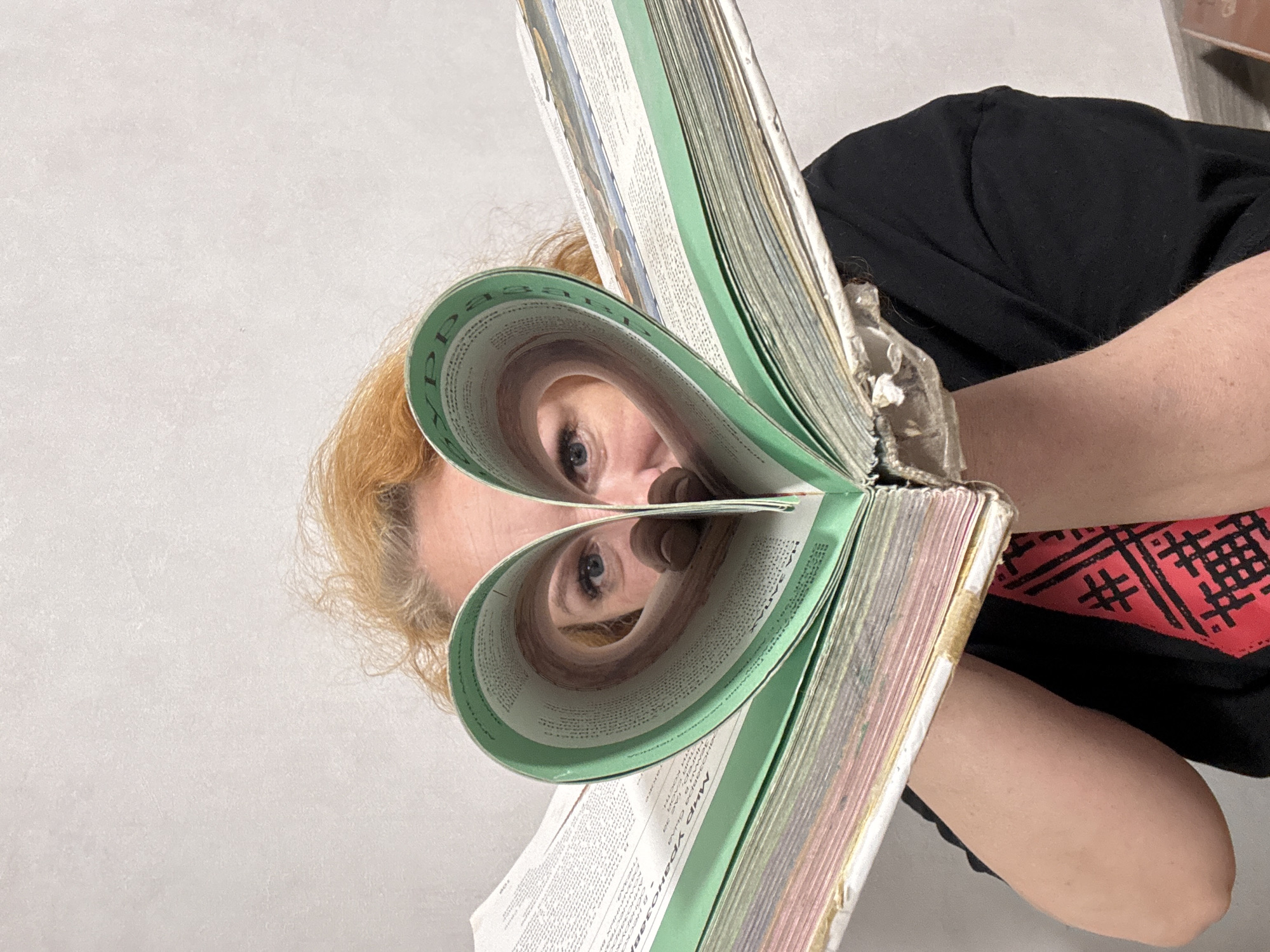 The image captures a close-up, creative portrait of a person, likely a woman, against a plain light-colored wall. The subject's face is partially obscured by a very thick book or directory, the pages of which have been skillfully bent and folded to create two distinct heart shapes. The person's blue eyes are visible peering through the openings of these heart-shaped folds, creating an illusion of eyes framed by hearts.
The subject has reddish-blonde, somewhat curly hair that frames her face. She is wearing a dark, short-sleeved top, possibly black, with a portion of a red fabric displaying a black geometric pattern visible on her right side. Her arm is also visible.
The book is substantial, appearing old and well-used, with numerous pages and distinctive green-colored edges on many sections. The spine or binding shows signs of wear, possibly mended with a gold or silver-colored material. Visible text on the pages is in Cyrillic script. One clearly discernible phrase is "МИР УРАВНОВЕШИВАЕТСЯ," which translates to "The world balances." Other partial words like "ррра" are also visible due to the page curvature.
The activity depicted is an artistic pose using the book to frame the eyes in heart shapes, suggesting playfulness or a romantic theme. The setting is indoors, with the light-colored wall providing a simple and unobtrusive background. The image was taken in Chaikovsky, Russia, at an indeterminate time of day.