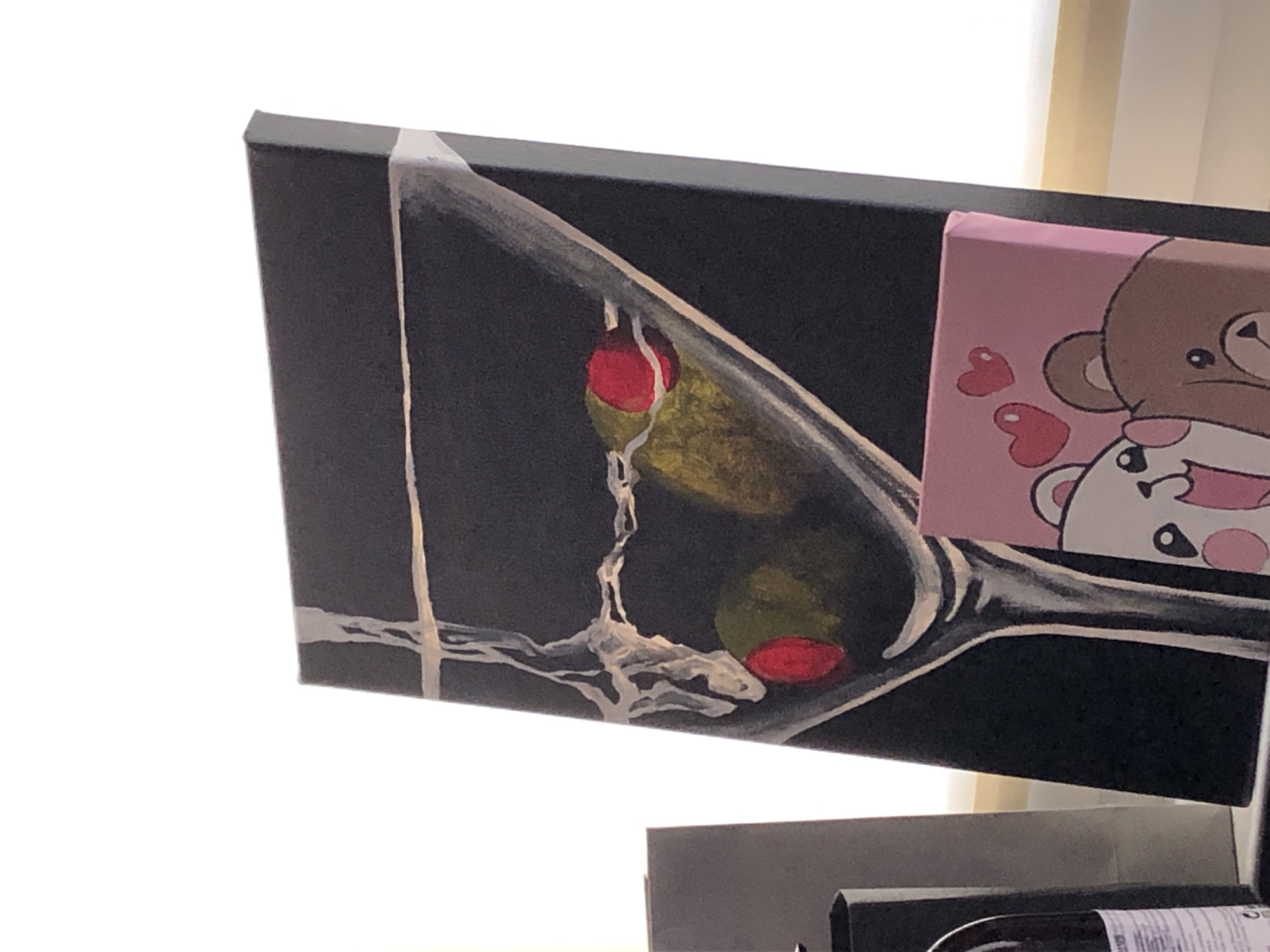 The image shows a close-up of a black canvas painting depicting a martini glass with olives. The olives have red pimento stuffing. Next to the martini painting is a smaller pink canvas with a cartoon drawing of two bears with hearts. Below these canvases, the top of a wine bottle is visible. The location and city are unknown.