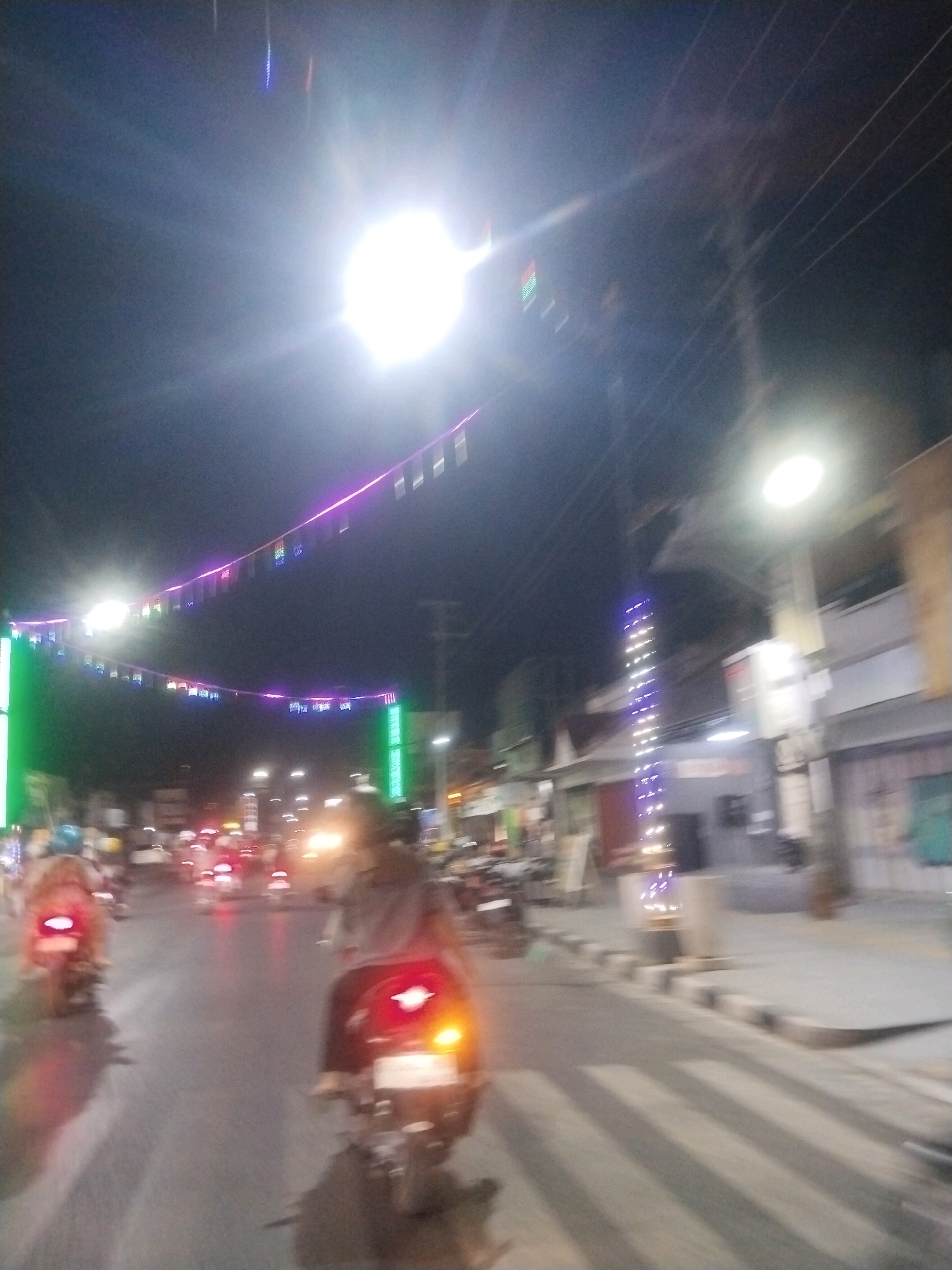 This is a low-light, motion-blurred photograph captured at night on a street in Pemalang, Indonesia. The scene depicts traffic on a road adorned with festive string lights that are suspended across the street. Several motorcycles are visible, with their taillights illuminated, indicating movement. Buildings line the side of the road, some featuring illuminated signage and decorative lights. The overall atmosphere suggests a vibrant street scene, possibly during a local celebration or event. The bright overhead lights and the lights from the buildings create a dynamic, illuminated urban environment despite the darkness of the night.