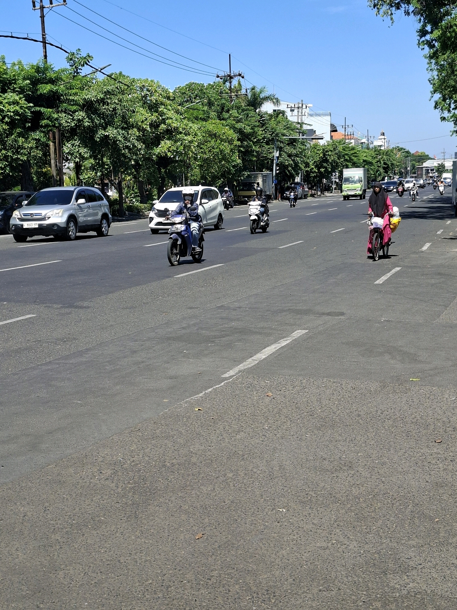 The image depicts a street scene in Surabaya, Indonesia, under a clear blue sky. The road is asphalt, with white lane markings. On the left side of the road, a silver SUV is parked. Further ahead, a white MPV is in motion, followed by a person on a blue motorcycle. A woman in a pink outfit and black hijab is riding a bicycle with a basket in front, also moving forward on the road. Numerous other vehicles, including motorcycles and cars, are visible further down the street, indicating moderate traffic. Trees with dense green foliage line the left side of the road, and buildings, some with white facades and others with visible rooftops, are present in the background. Power lines are suspended overhead. The overall impression is a typical urban street on a sunny day.