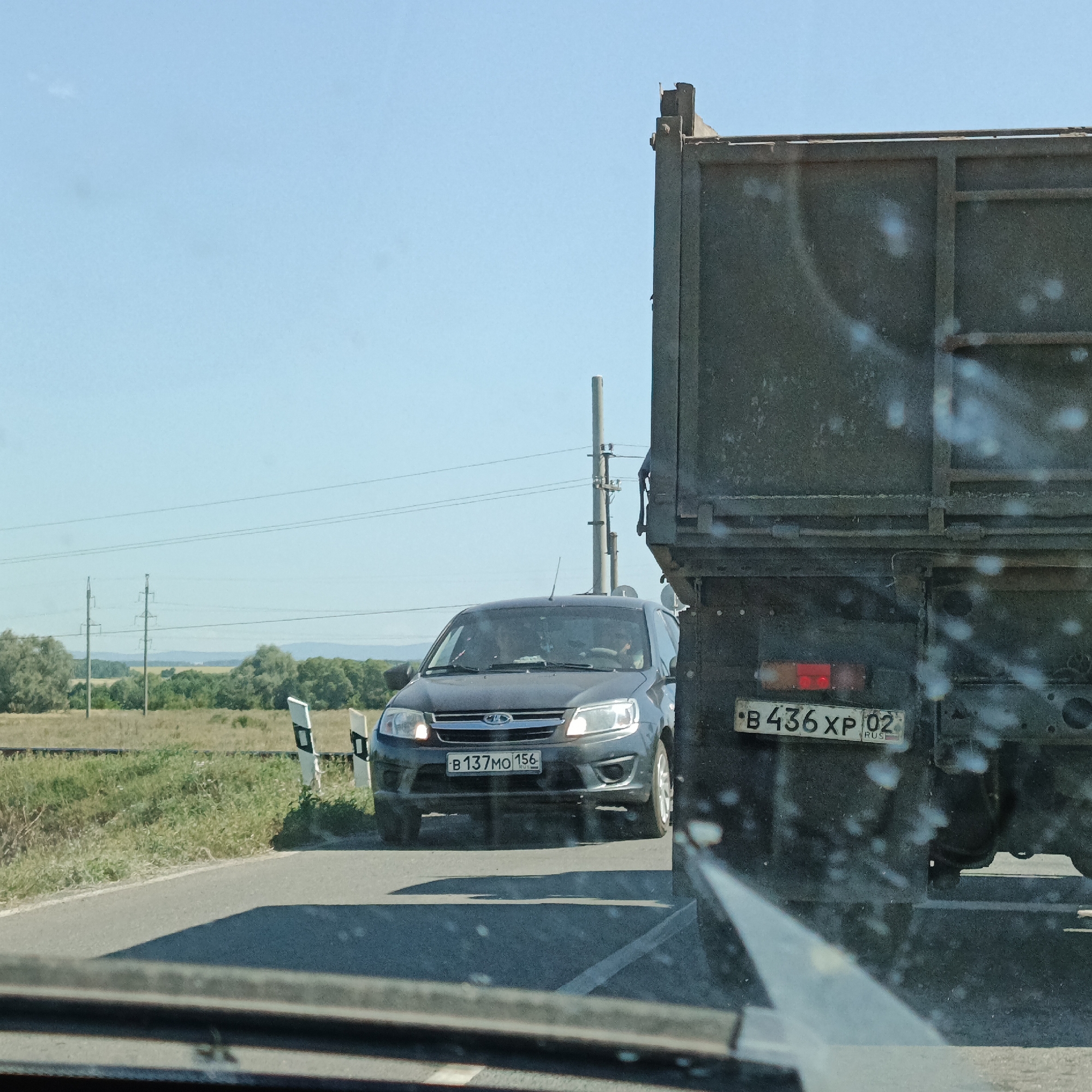 In Aksarovo, Russia, on a sunny day, a dark-colored sedan with the license plate "B 137MO 156" is stopped on a paved road. Behind the sedan, a large dark-colored truck, partially obscuring the view, is also stopped. The truck has a license plate that reads "B 436 XP 02". The environment includes a grassy embankment with vegetation and trees in the background, and utility poles with overhead wires are visible. The scene appears to be on the outskirts of town or in a rural area, with no other vehicles or pedestrians immediately present. The image seems to be taken from inside another vehicle, as indicated by the reflection on the windshield in the foreground.